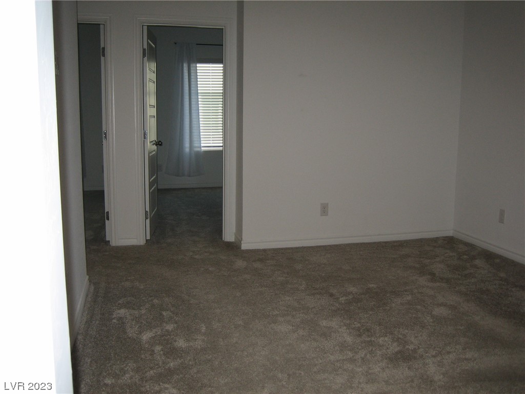 Empty carpeted room with neutral walls and an open doorway to a windowed room.