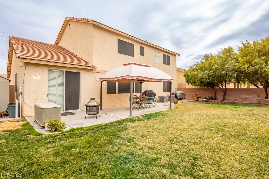 Two-story beige house with a grassy backyard, shaded patio dining set, and mature trees.