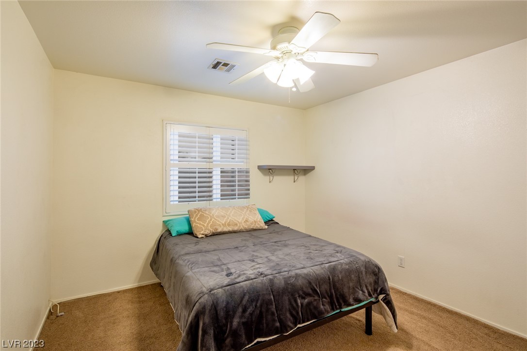 Bedroom with a dark gray-covered bed, shuttered window, tan carpet, and ceiling fan.
