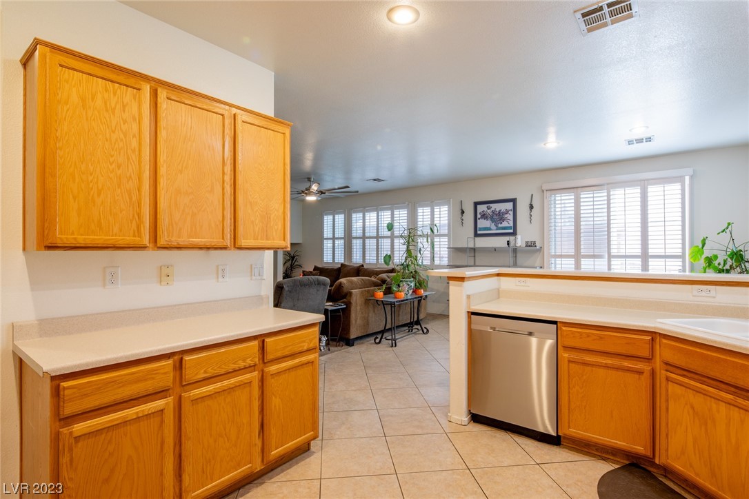 Open kitchen with honey-colored cabinets, stainless steel dishwasher, and a view into the furnished living room.