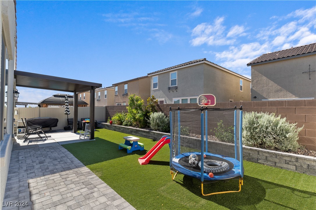 Backyard with artificial turf, covered patio, trampoline, and children's slide.