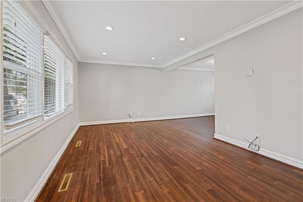 Empty room with dark wood flooring, wide windows with blinds, recessed lights, and crown molding.