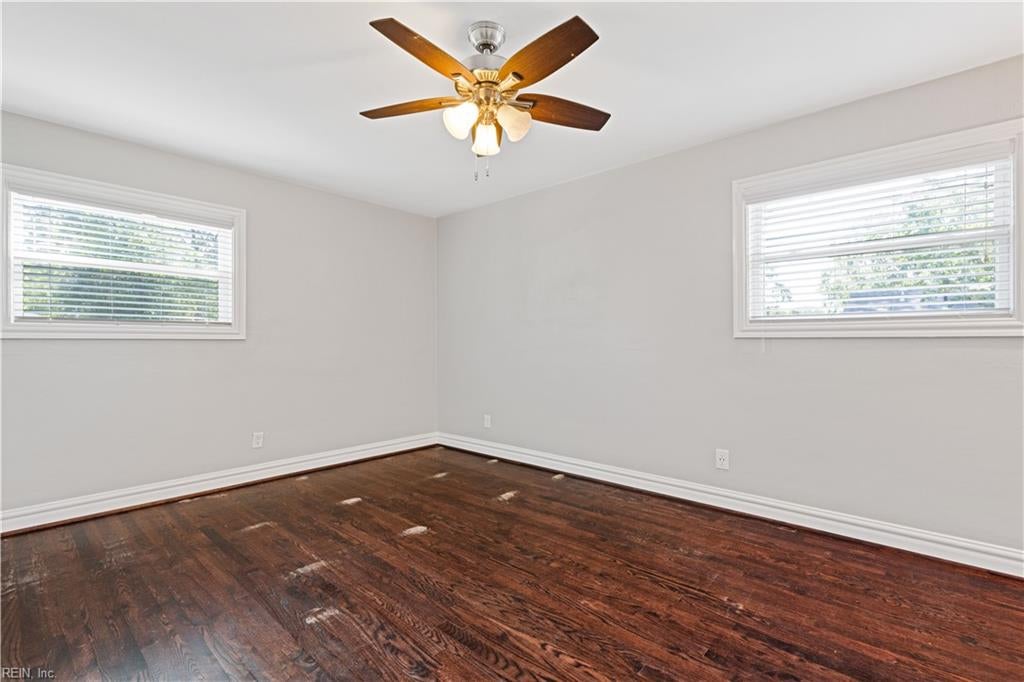 Empty room with dark hardwood floors, two windows with blinds, and a lit ceiling fan.