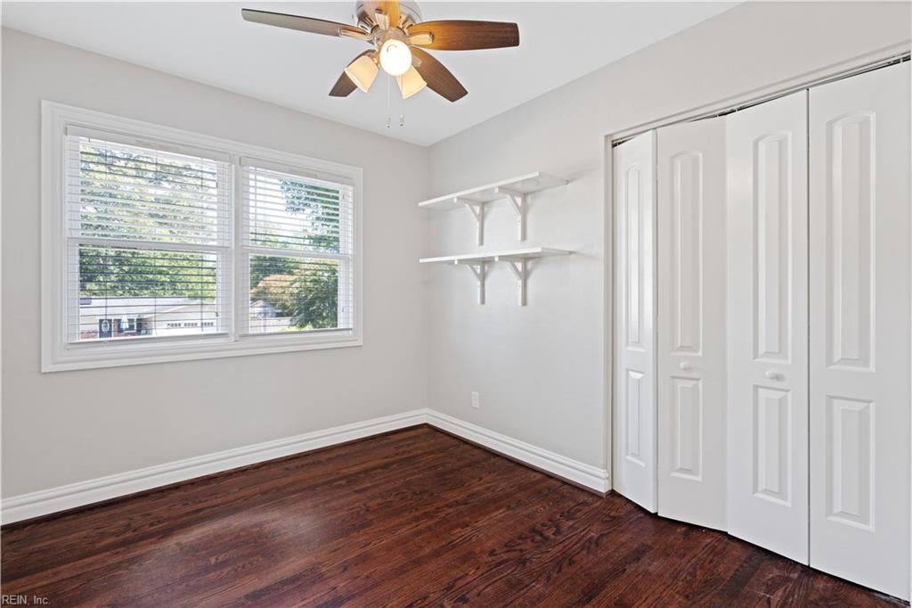 Empty bedroom with dark hardwood floors, a ceiling fan, open shelves, and folding closet doors.