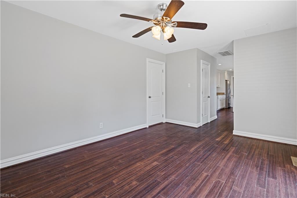 Vacant room with dark wood floors and a ceiling fan, opening toward the kitchen.