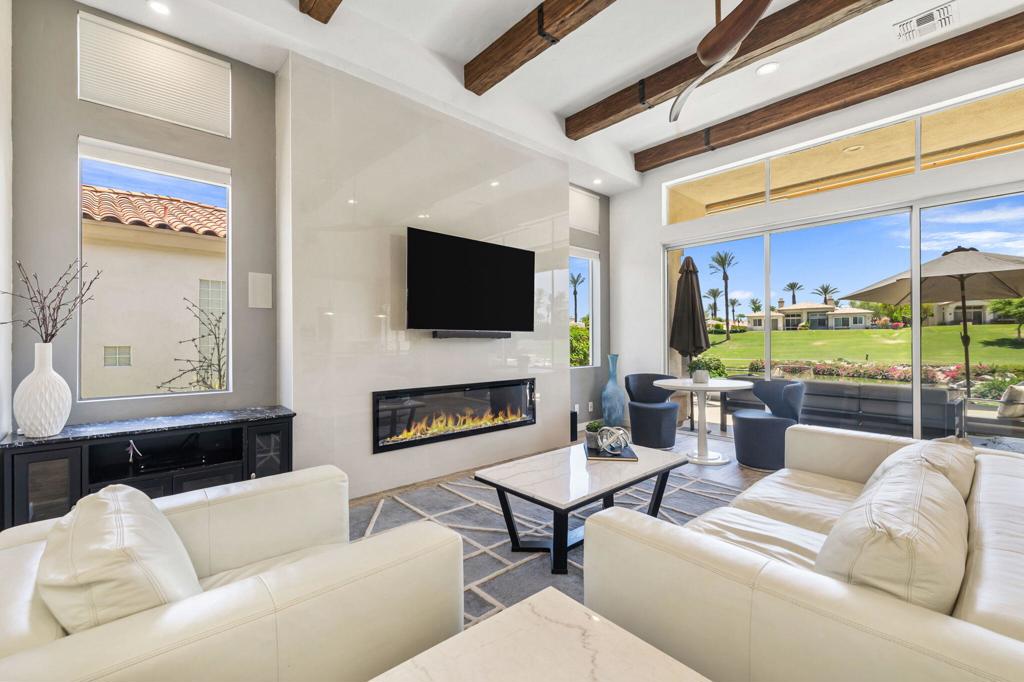 Living room with white leather sofas, a linear fireplace, exposed wood beams, and views of a green lawn through glass doors.