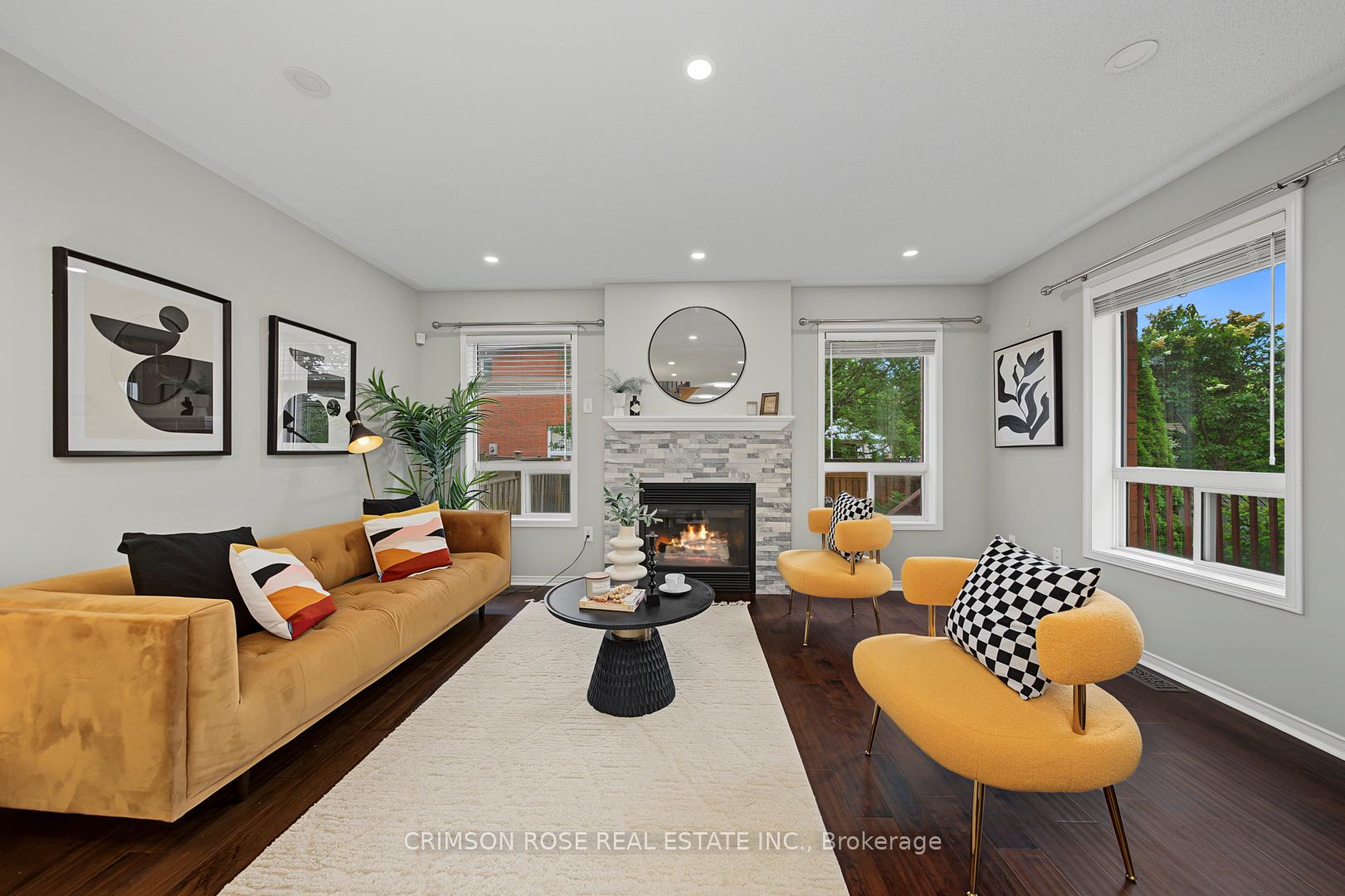 Staged living room with a stone fireplace, mustard-yellow seating, dark hardwood floors, and large windows.