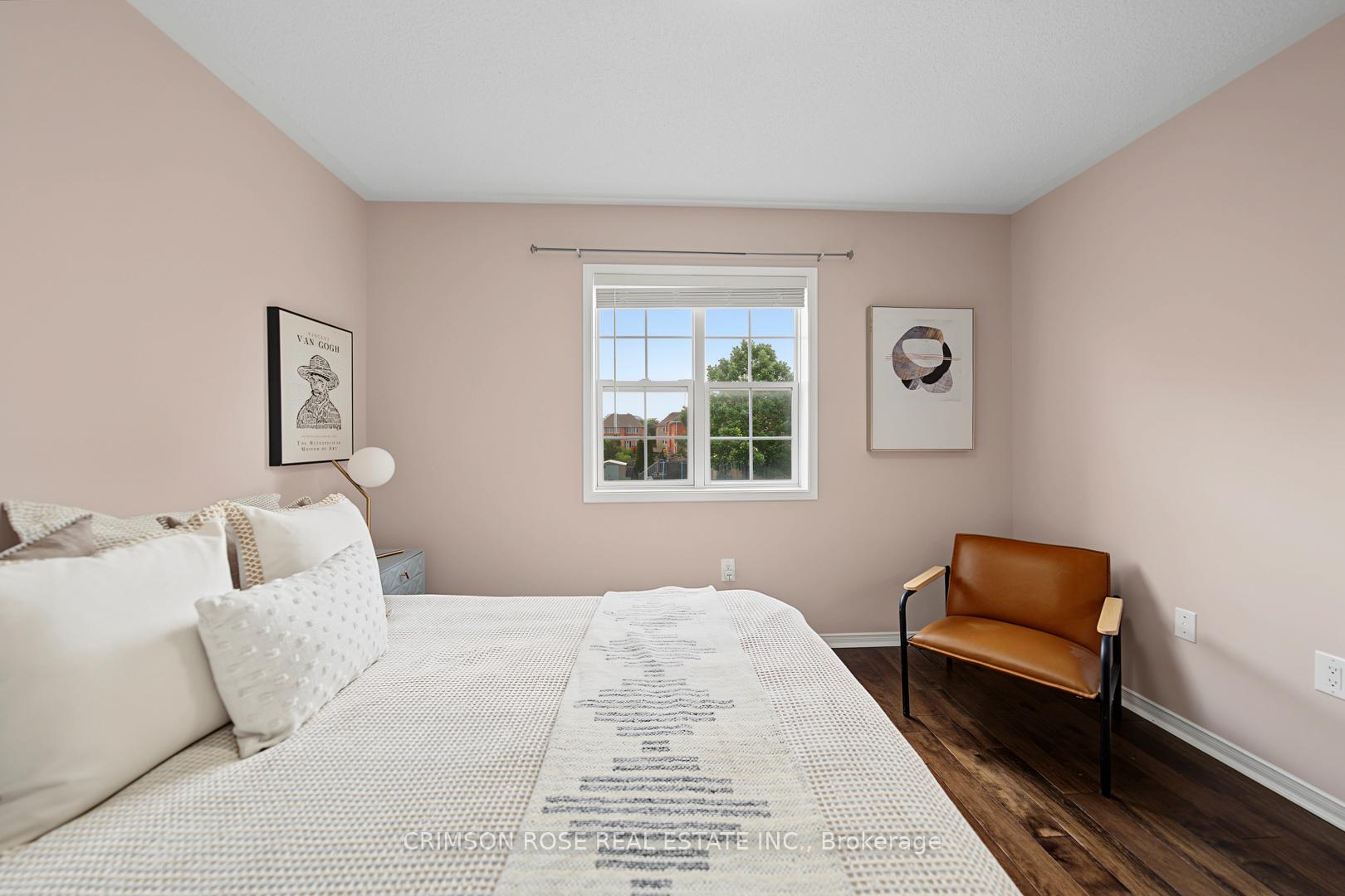Furnished bedroom with a white bed, brown leather chair, and window overlooking trees and neighboring homes.
