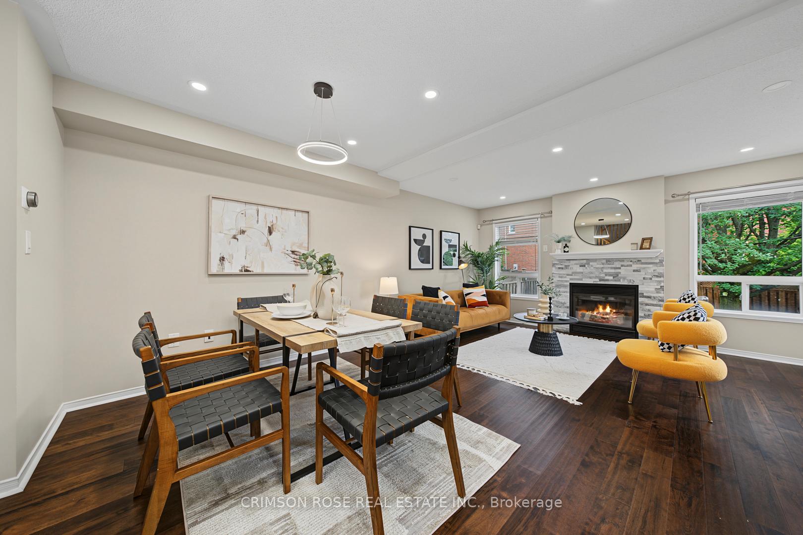 Open-plan living and dining room with a stone fireplace, dark hardwood floors, and windows overlooking greenery.