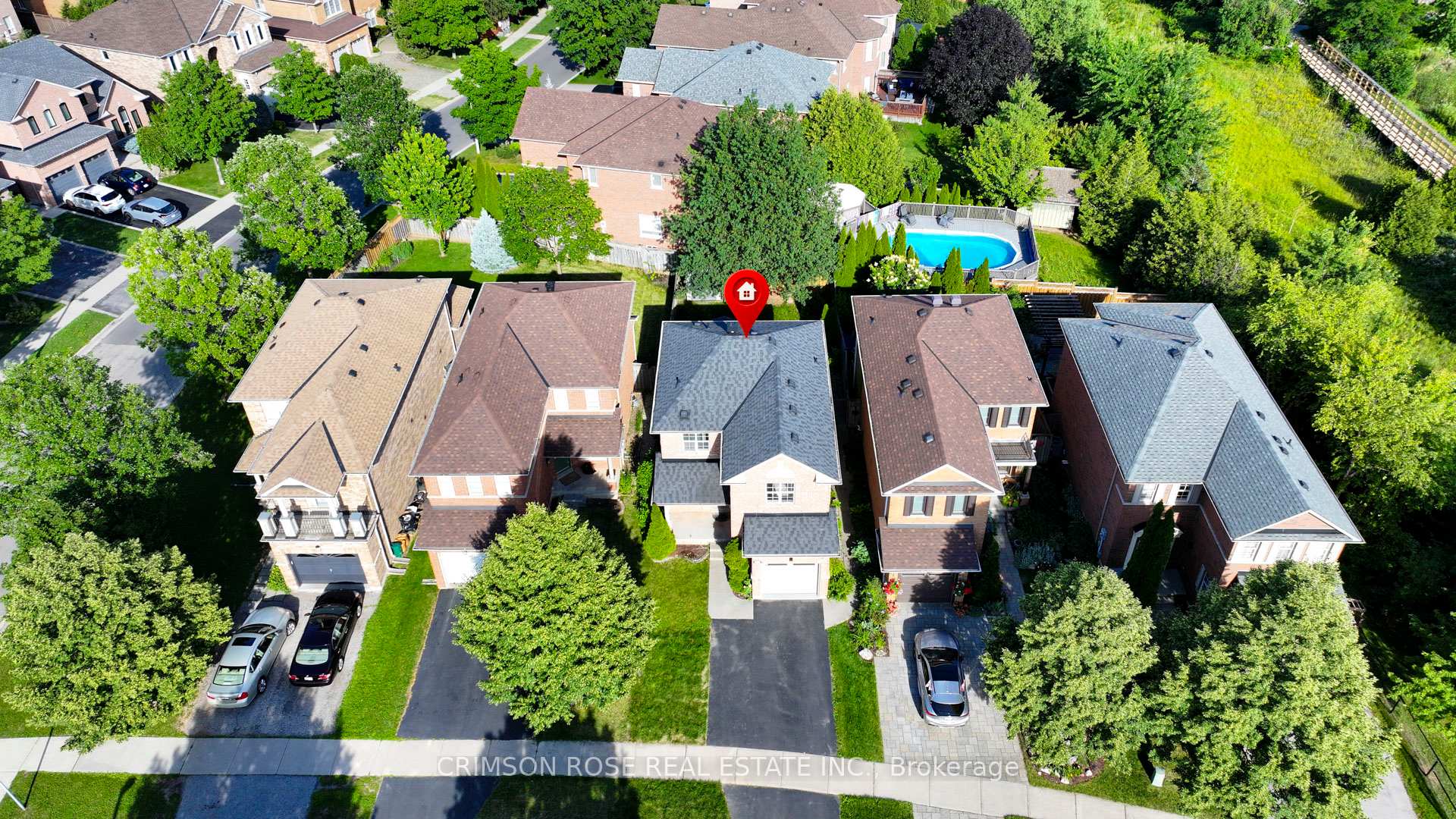 Aerial view of a detached brick house with a gray roof, driveway, and landscaped yard among leafy neighboring homes.