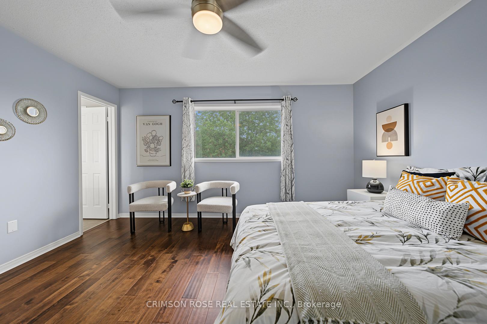 Furnished bedroom with a large patterned bed, hardwood floors, and two chairs beside a window.