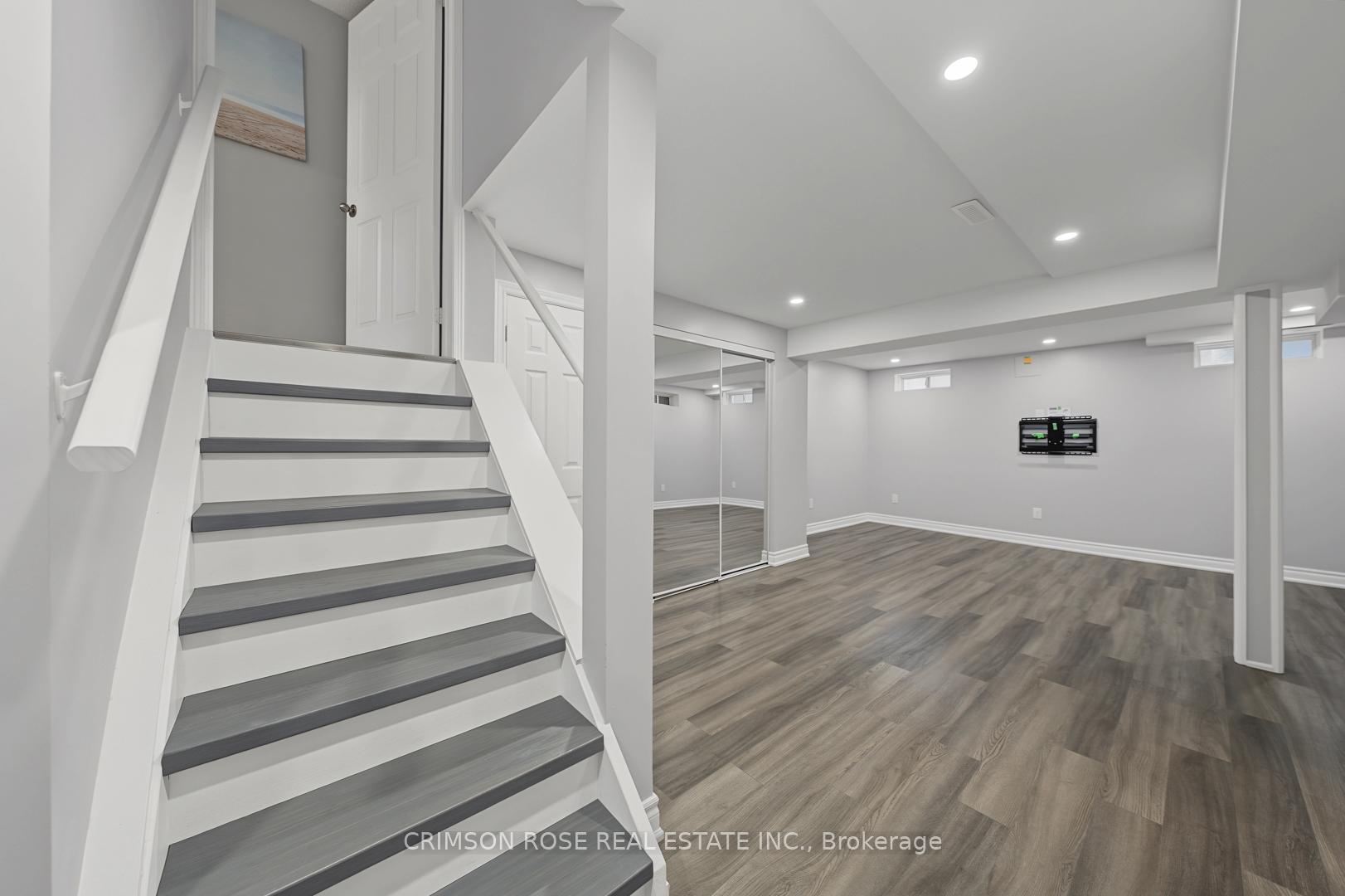 Basement recreation room with gray flooring, mirrored closet doors, and a staircase with dark treads.