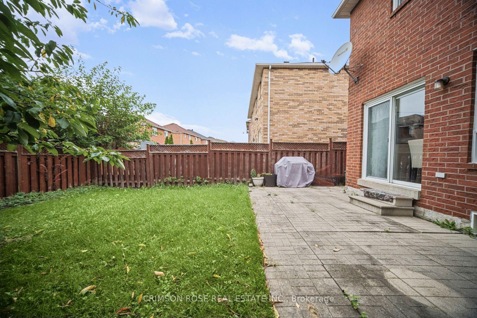 Fenced backyard with a grassy lawn, paved patio, and covered grill beside a red-brick house.