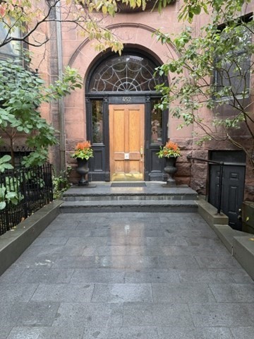 Brick building entrance with an arched doorway, wooden door, stone walkway, and potted plants.
