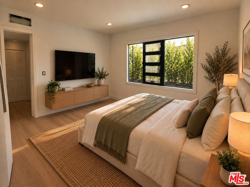 Furnished bedroom with a large neutral bed, woven rug, and wide window overlooking greenery.