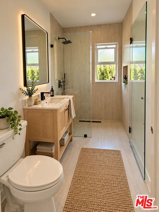 Modern bathroom with a glass walk-in shower, light wood vanity, and window overlooking greenery.