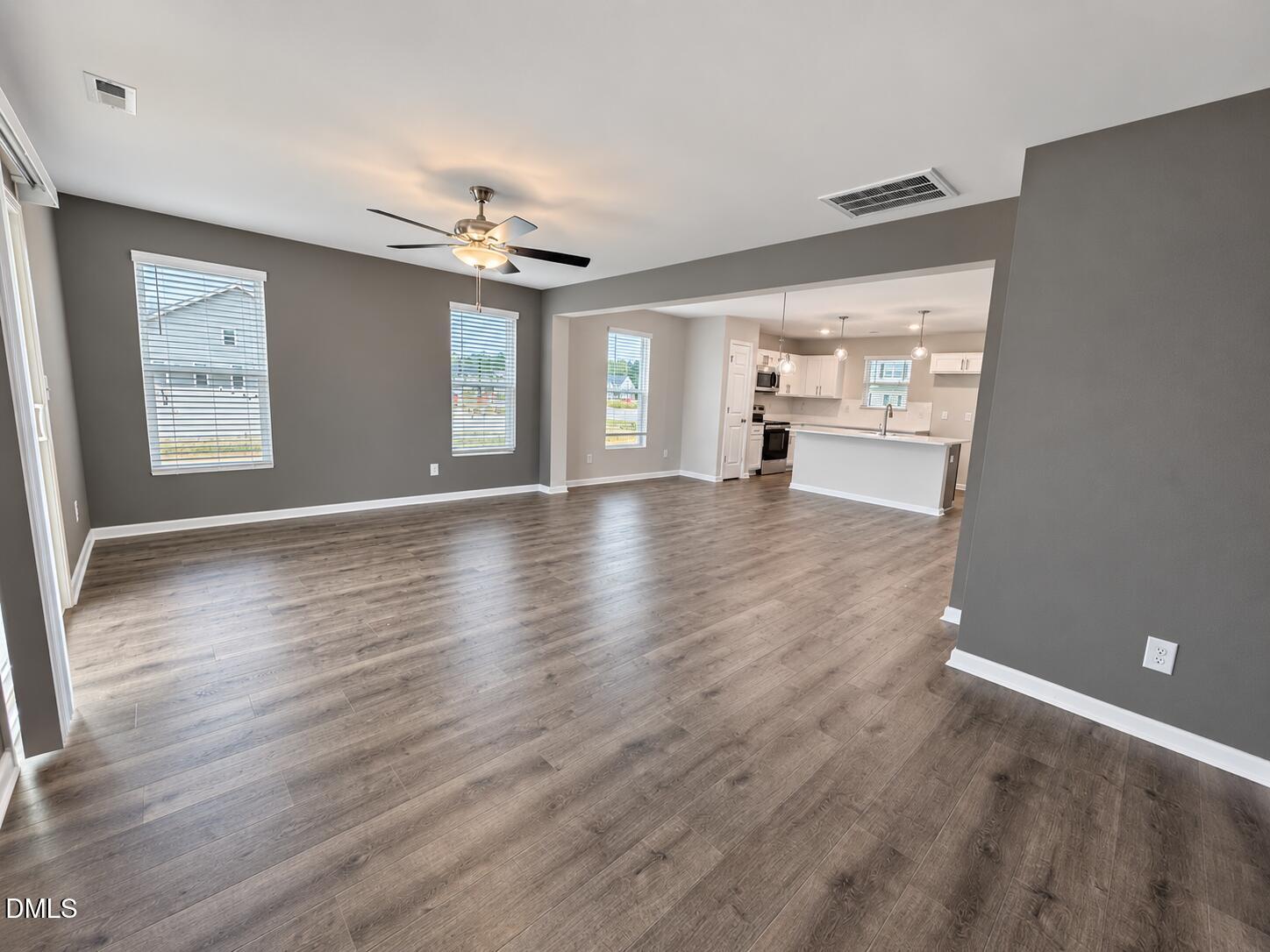 Empty living room with wood-look floors and ceiling fan, opening to a white kitchen with an island.
