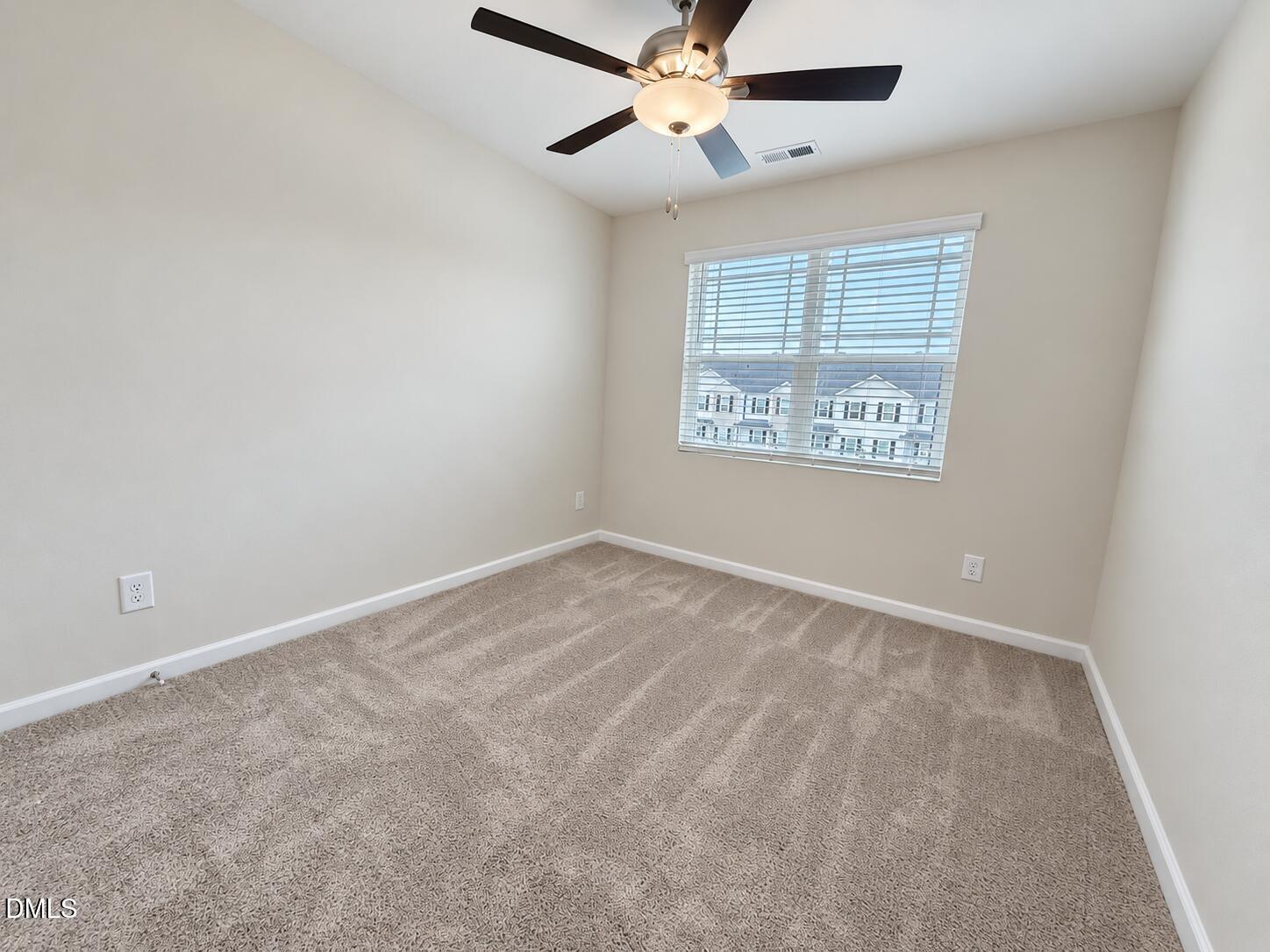 Empty carpeted bedroom with a ceiling fan and a window overlooking neighboring homes.