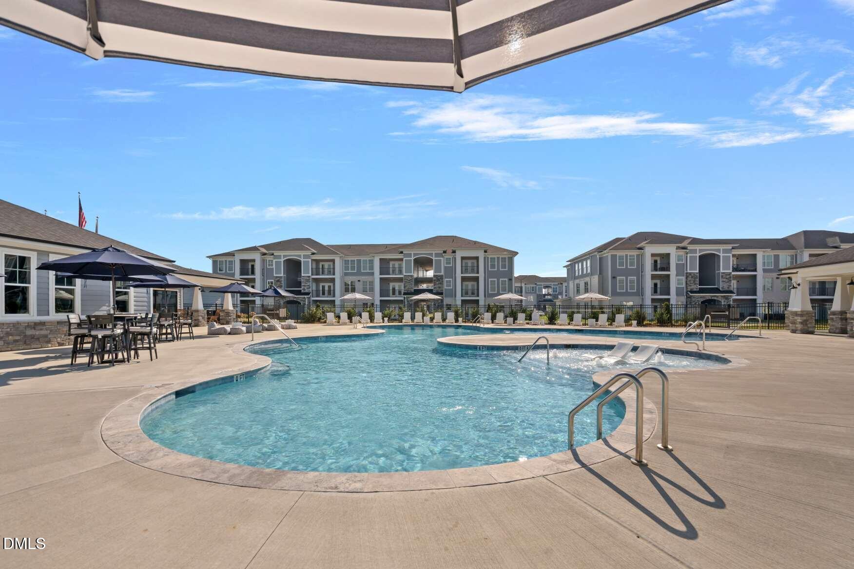 Community swimming pool with submerged lounge chairs, patio umbrellas, and apartment buildings.
