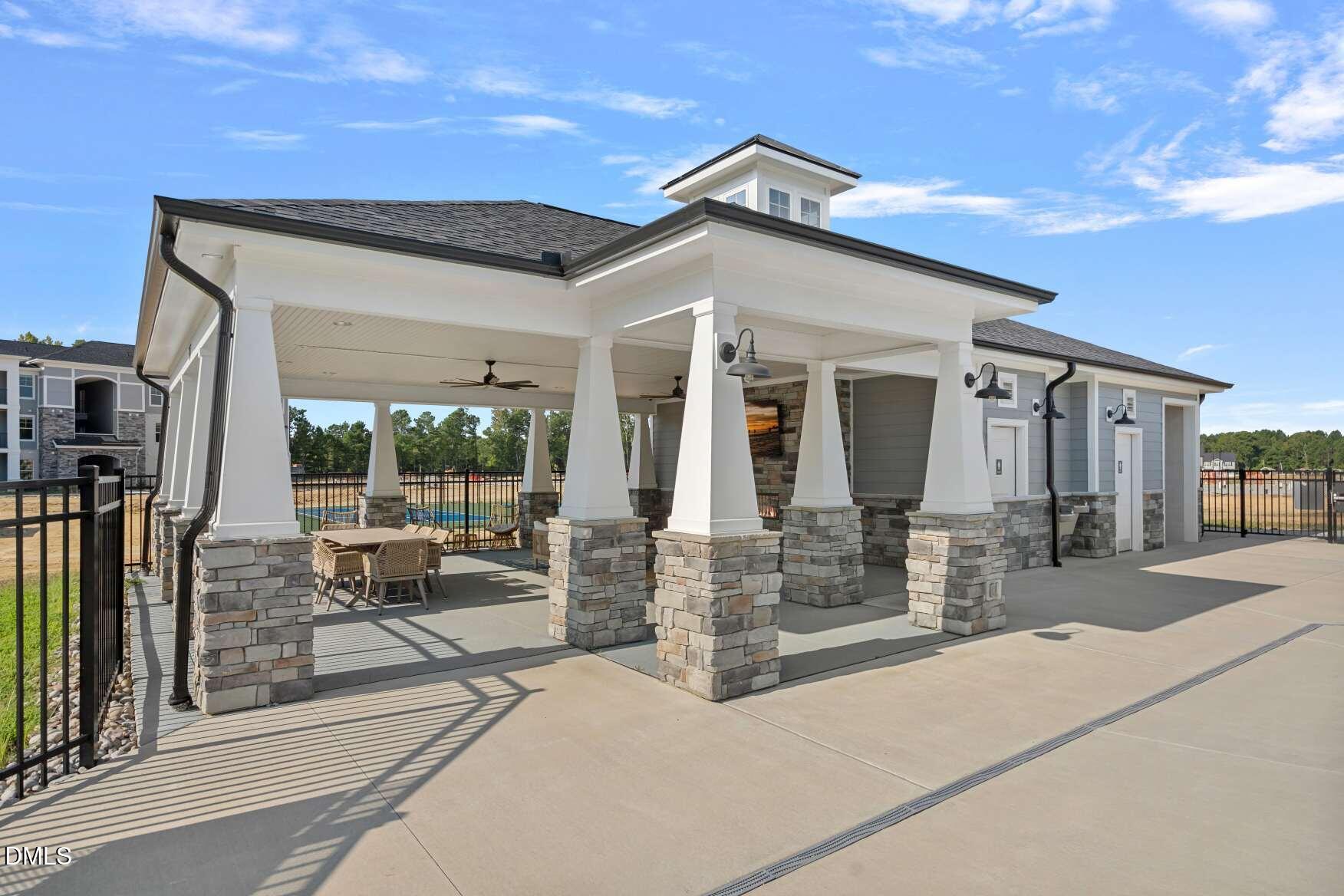 Covered community pavilion with stone columns, patio dining furniture, and a swimming pool beyond the fence.