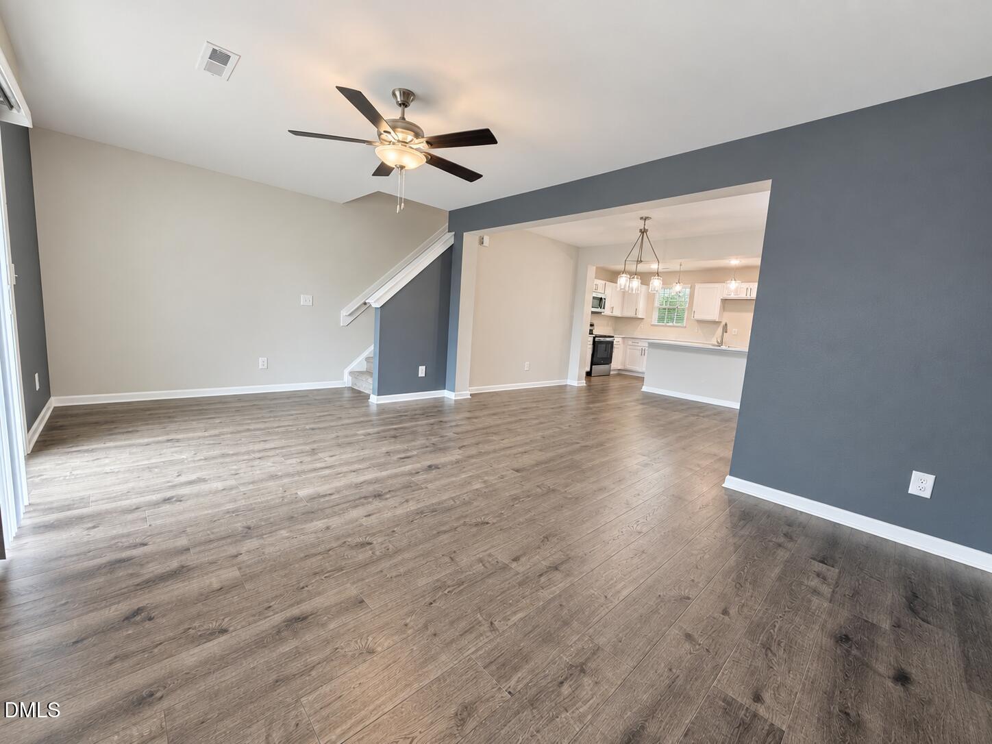 Empty living room with wood-look floors and ceiling fan, opening into a white kitchen with a breakfast bar.