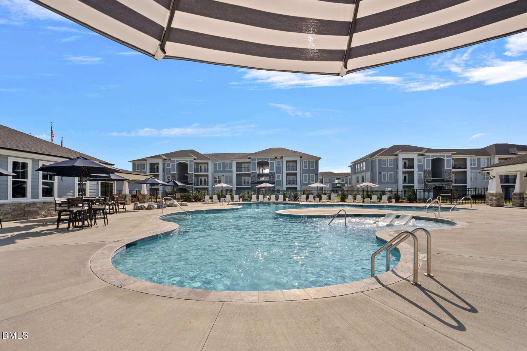 Apartment community pool with a curved swimming area, patio umbrellas, and multistory buildings.
