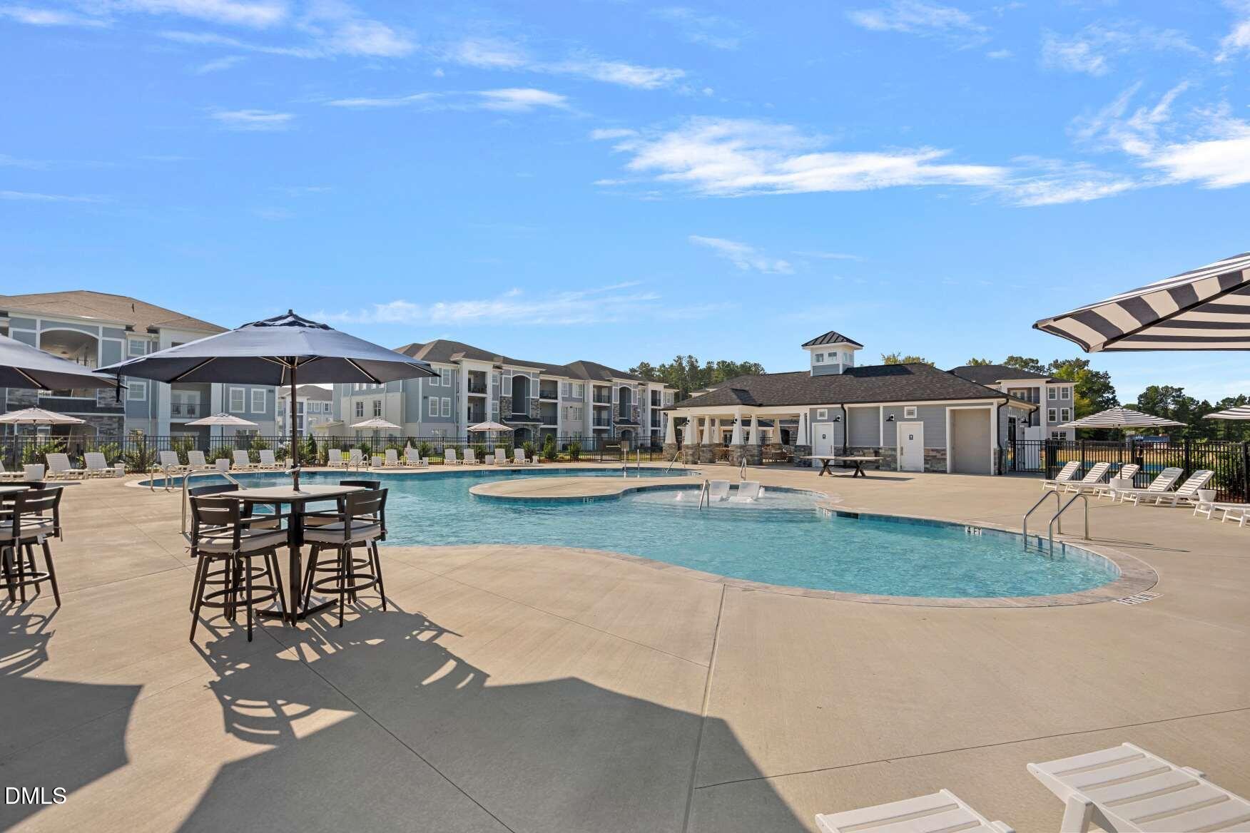 Community swimming pool with a sun deck, patio seating, umbrellas, and residential buildings.