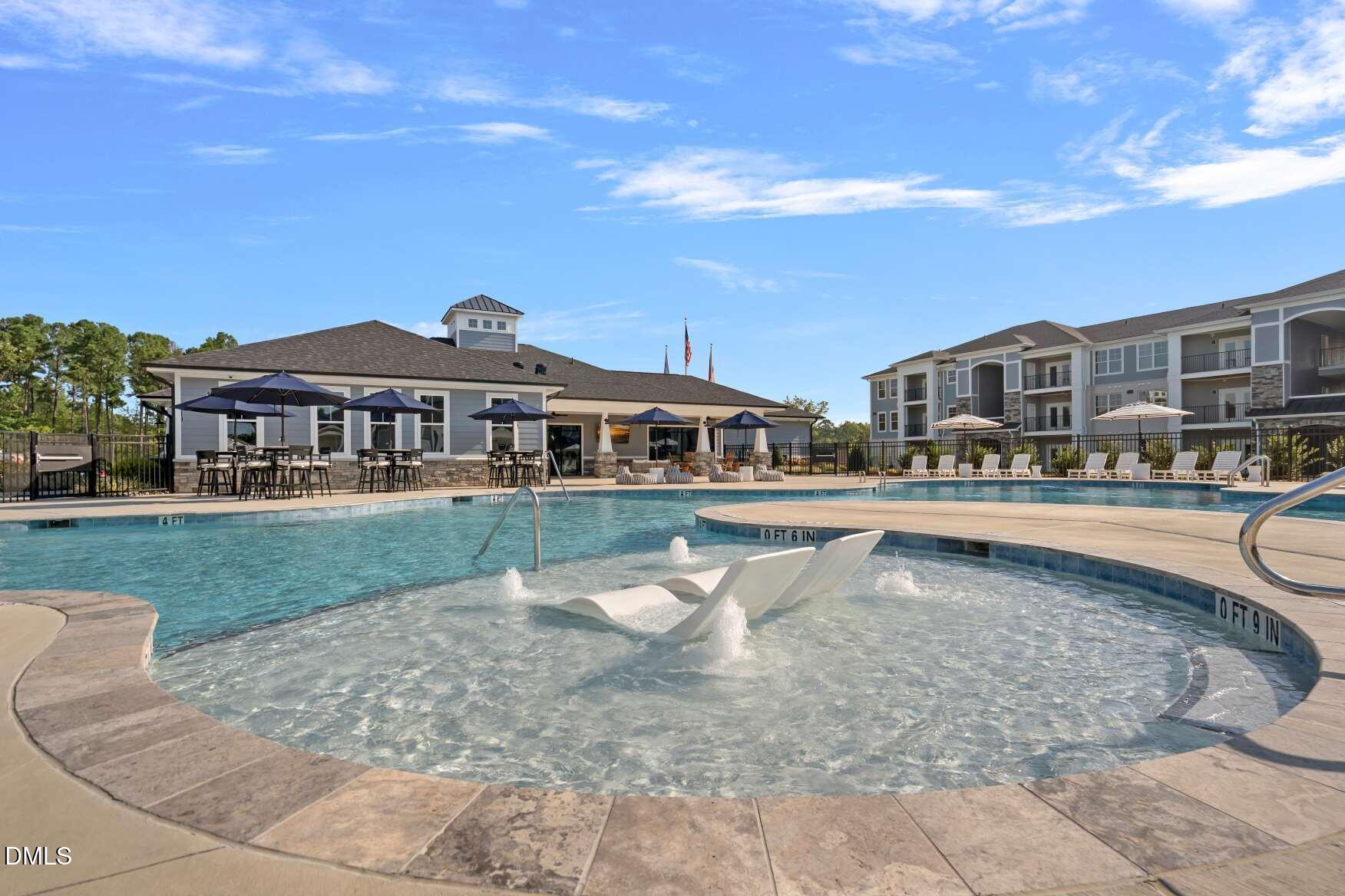 Community pool with a shallow sun shelf, in-water loungers, and apartment buildings beyond.