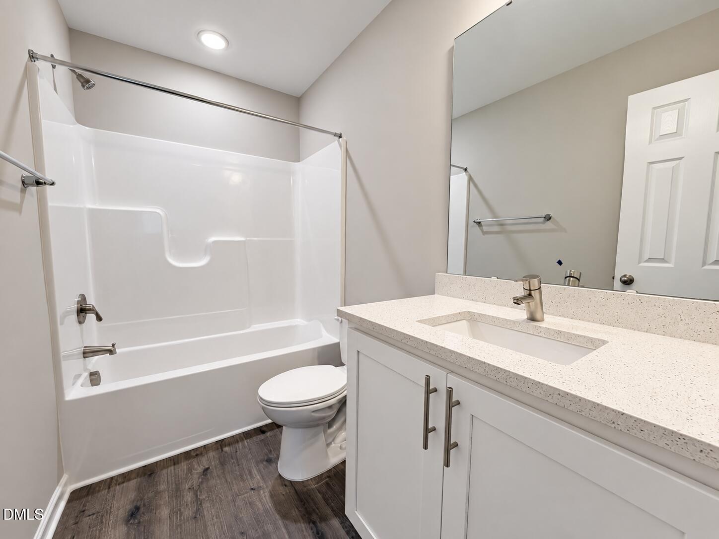 Bathroom with a white tub and shower, toilet, and speckled countertop vanity over dark wood-look flooring.