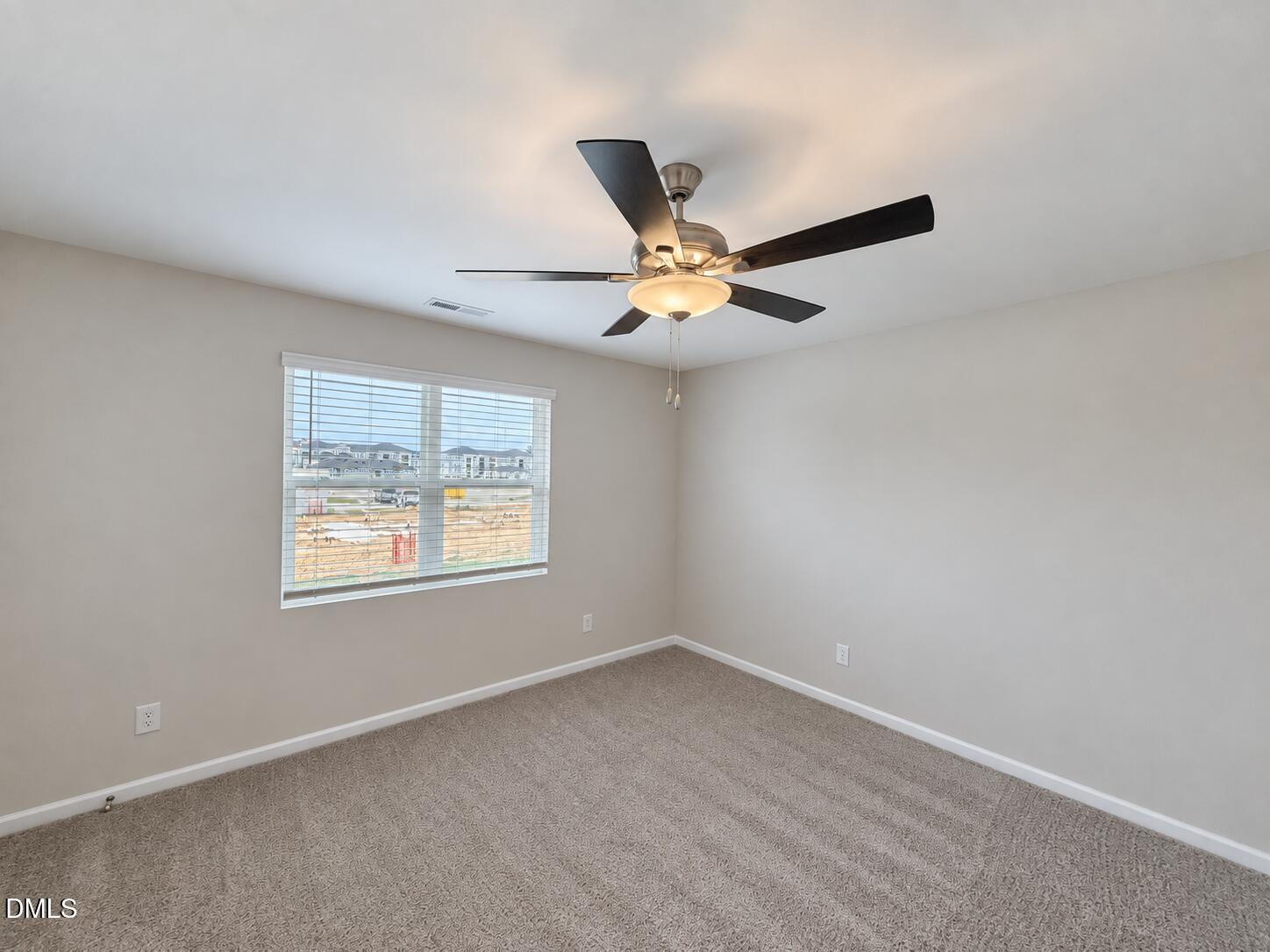 Empty bedroom with beige carpet, a dark-bladed ceiling fan, and a window overlooking nearby construction.