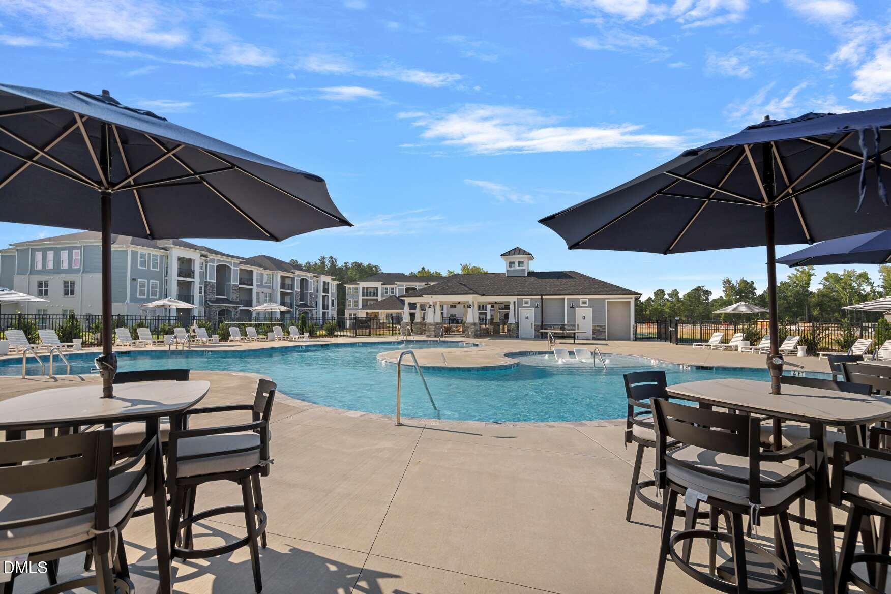 Community pool with a sun shelf, shaded patio tables, lounge chairs, and residential buildings.