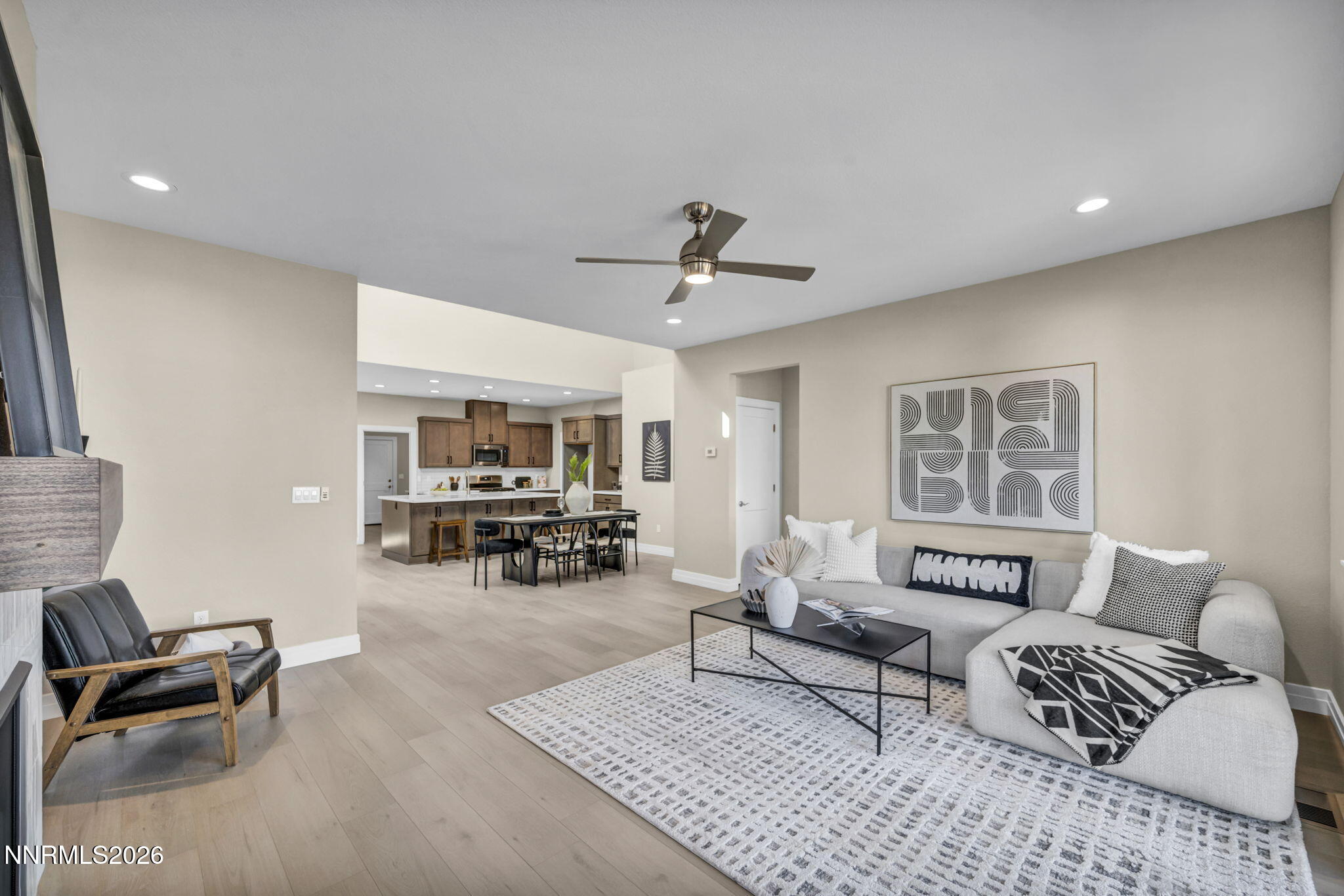 Staged living room opens to a dining area and kitchen, with a gray sectional and light wood floors.