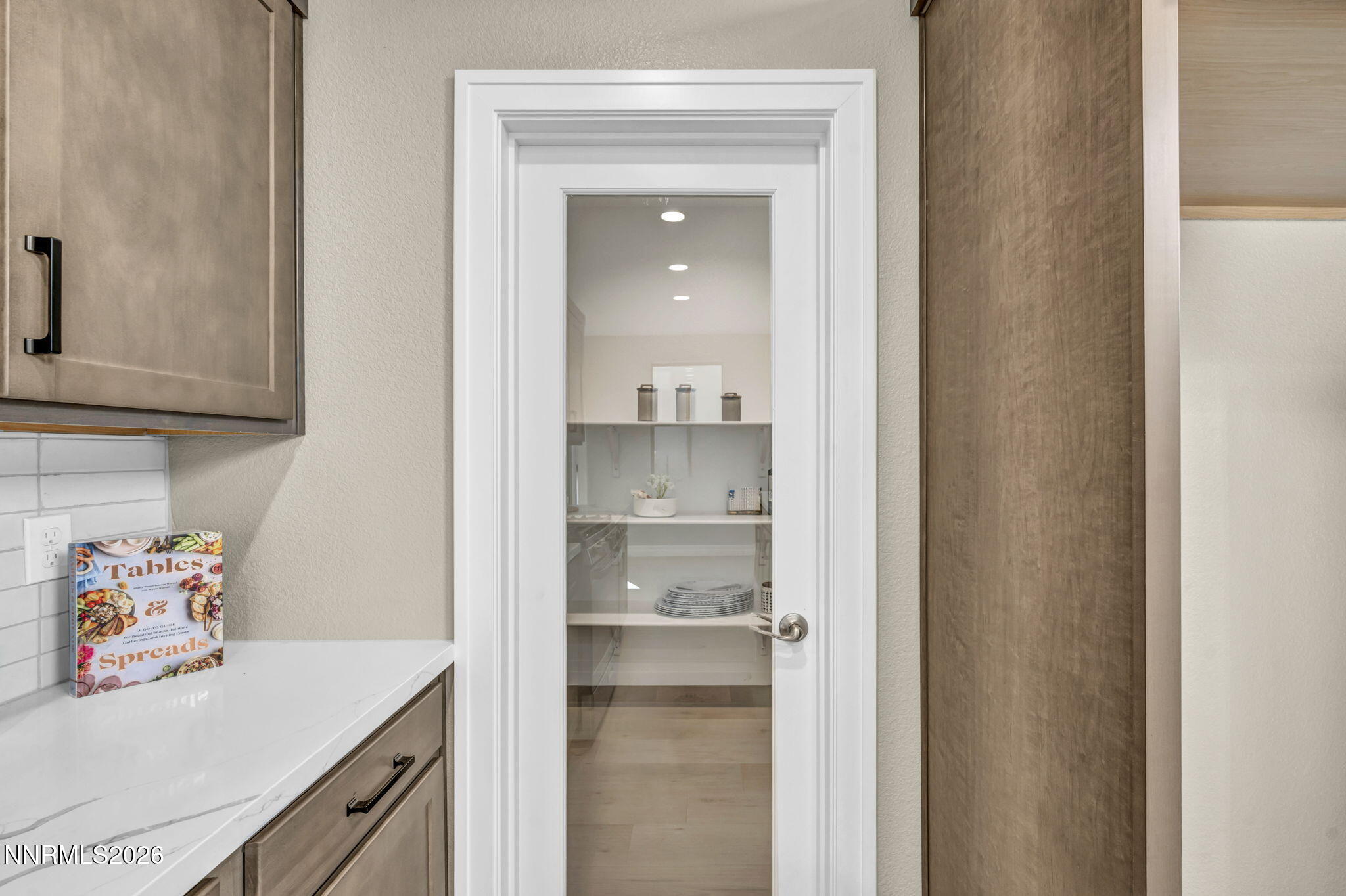Kitchen counter and taupe cabinets beside a glass-panel pantry door with dishes on its shelves.
