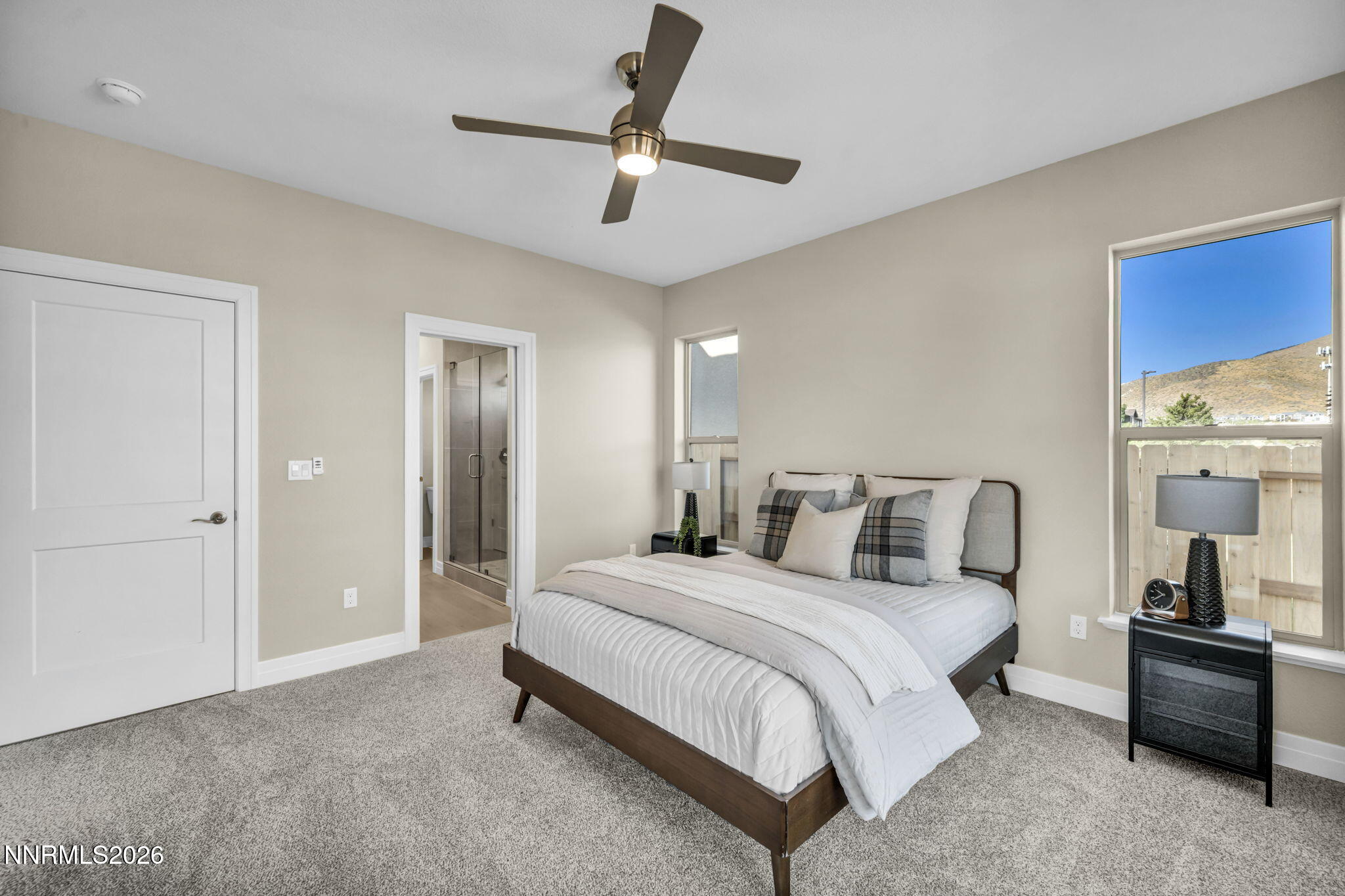 Staged bedroom with a white bed, ceiling fan, and window overlooking a dry hillside.