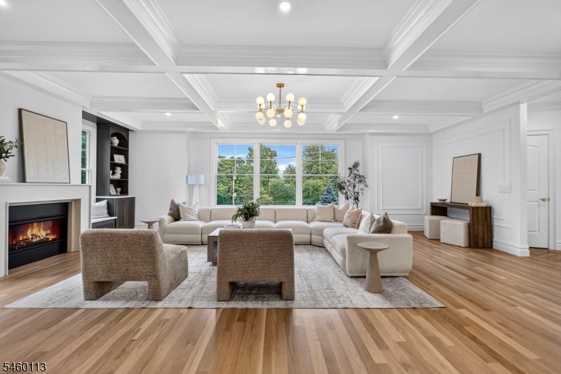 Staged living room with a cream sectional, fireplace, coffered ceiling, and large windows overlooking trees.
