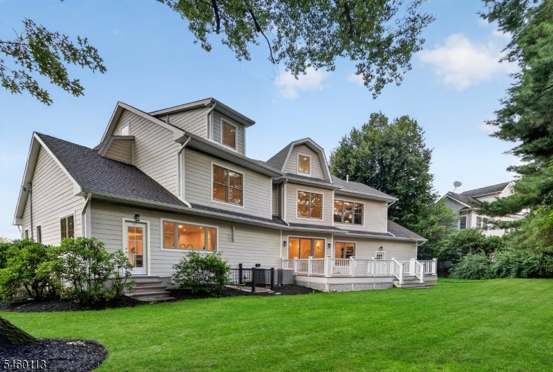 Gray multi-story house with a white-railed deck, illuminated windows, and a broad green lawn.