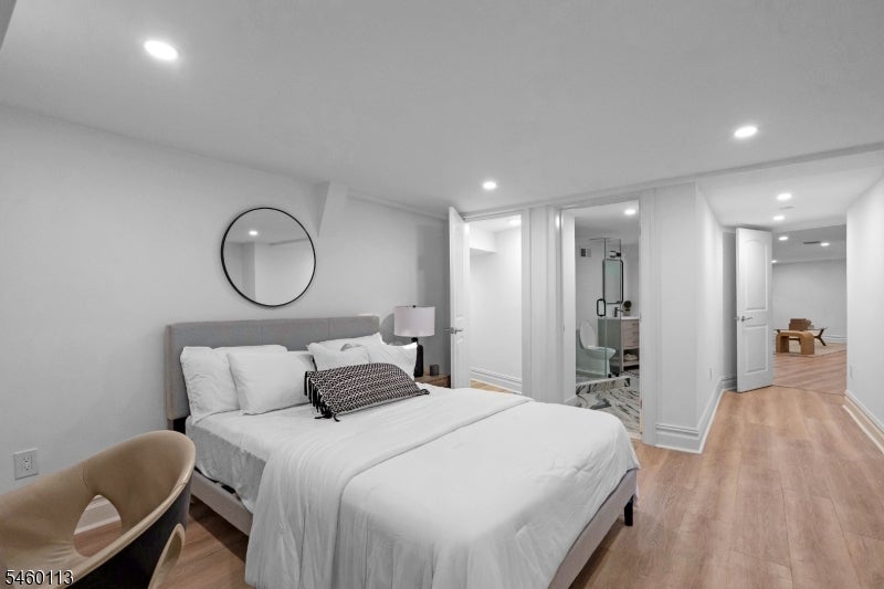 Staged bedroom with a gray upholstered bed, round mirror, ensuite bathroom, and light wood flooring.