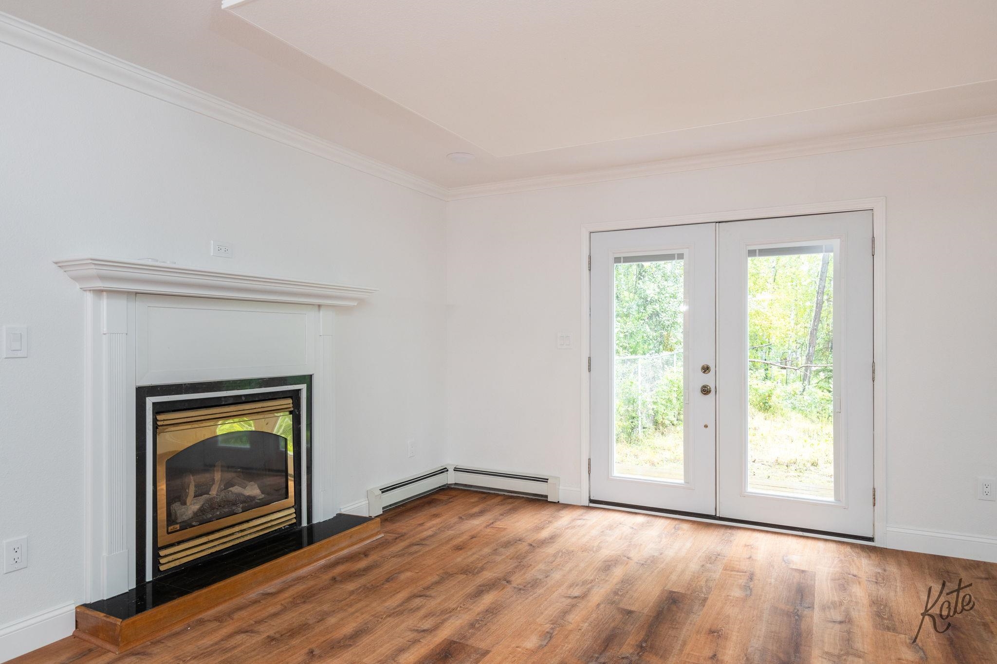 Empty living room with a white fireplace, wood floors, and double glass doors overlooking trees.