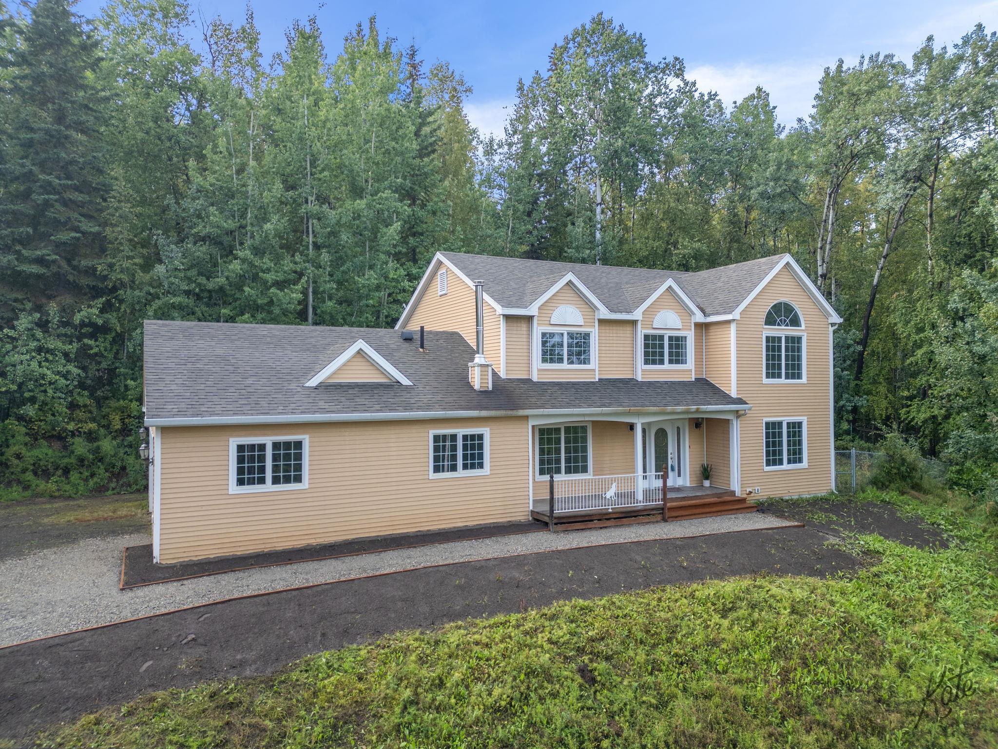 Tan two-story home with a covered porch, gravel drive, and wooded surroundings.