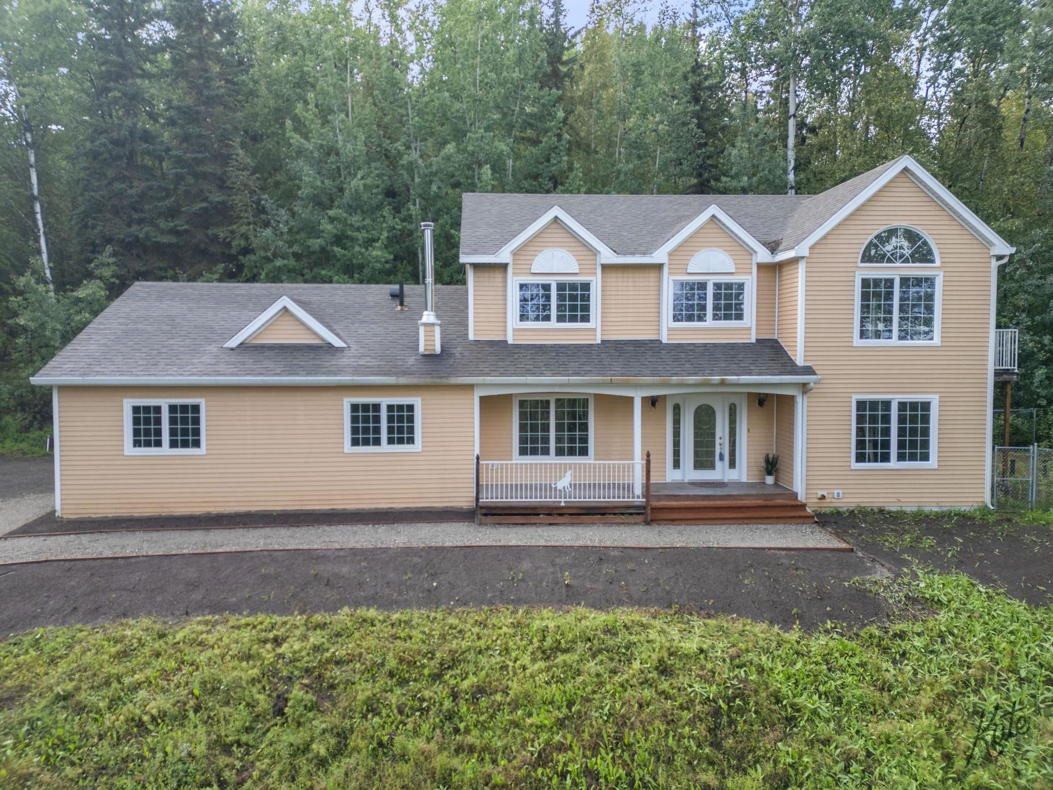 Tan two-story house with white trim, a covered porch, and dense trees behind it.
