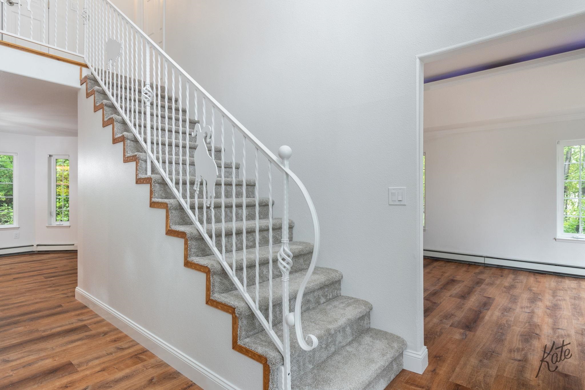 Carpeted staircase with white decorative railings and wood trim, between two rooms with wood-look flooring.