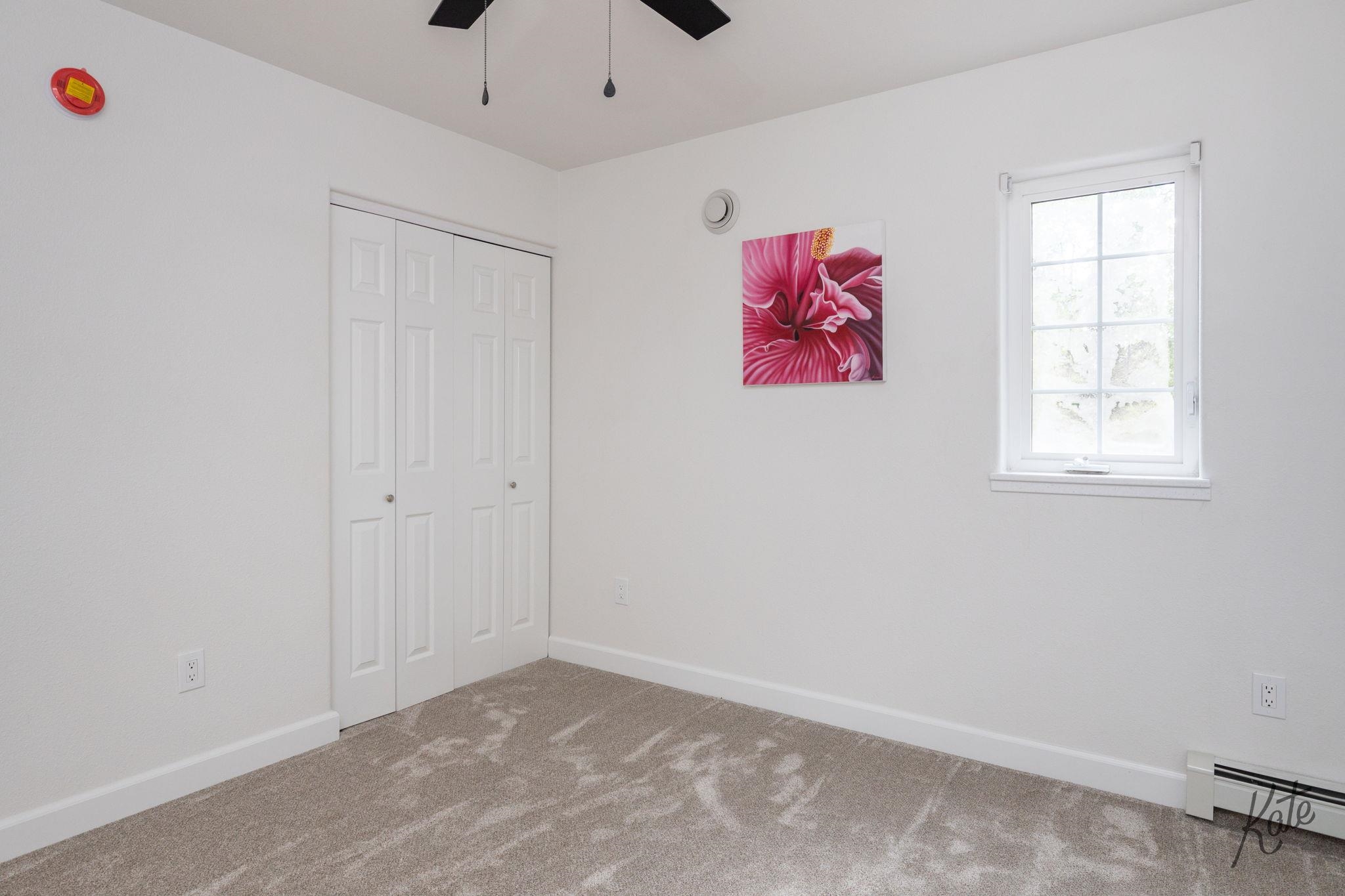 Empty bedroom with beige carpet, folding-door closet, bright window, and pink flower print.
