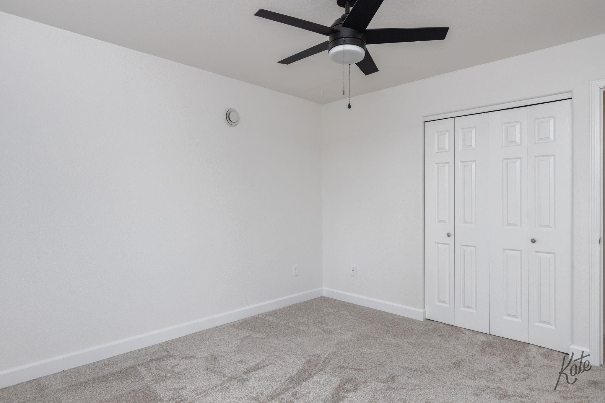 Empty carpeted bedroom with a black ceiling fan and white folding closet doors.