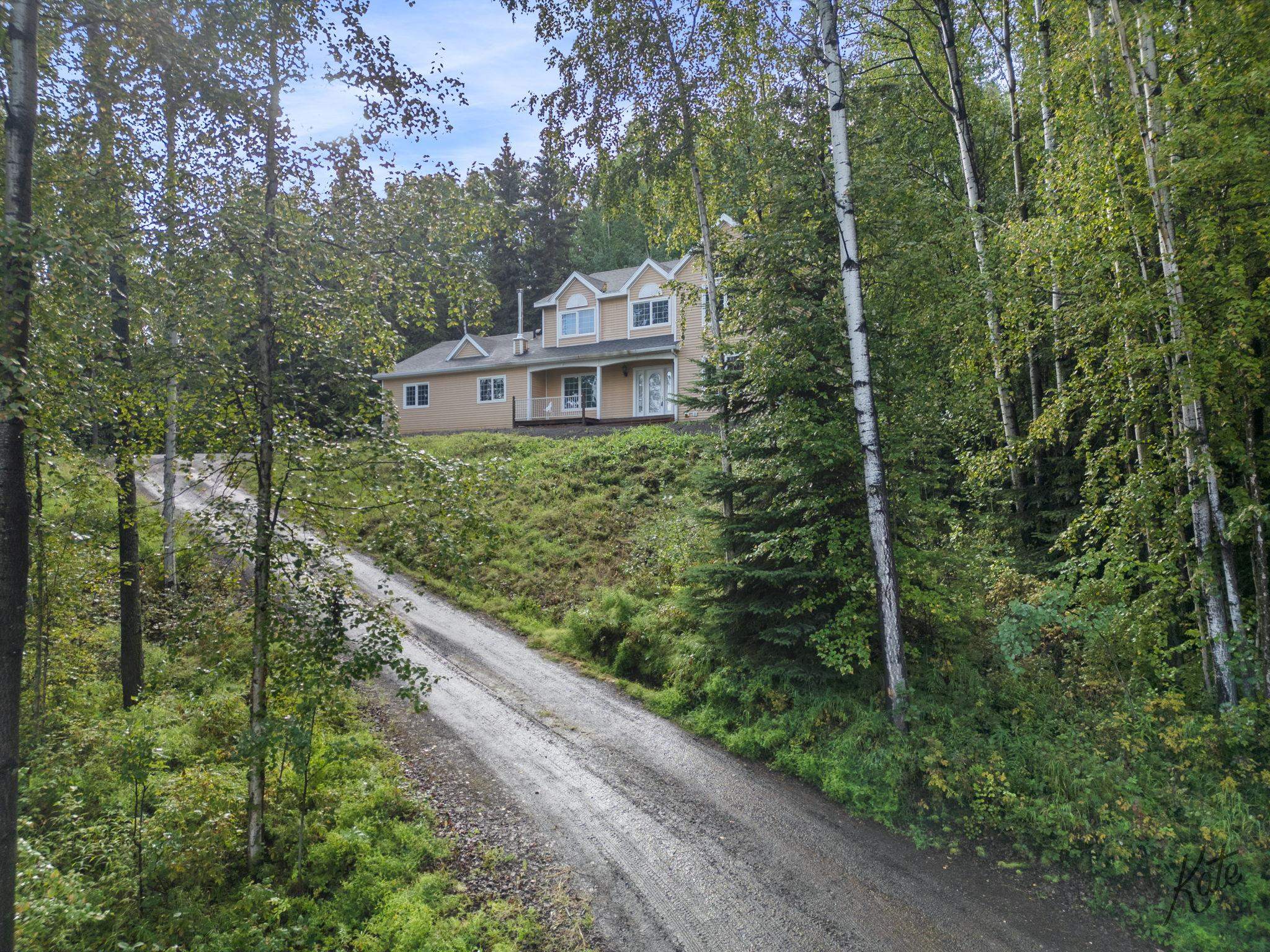 Tan two-story home with a covered porch, reached by a curving gravel driveway through dense trees.