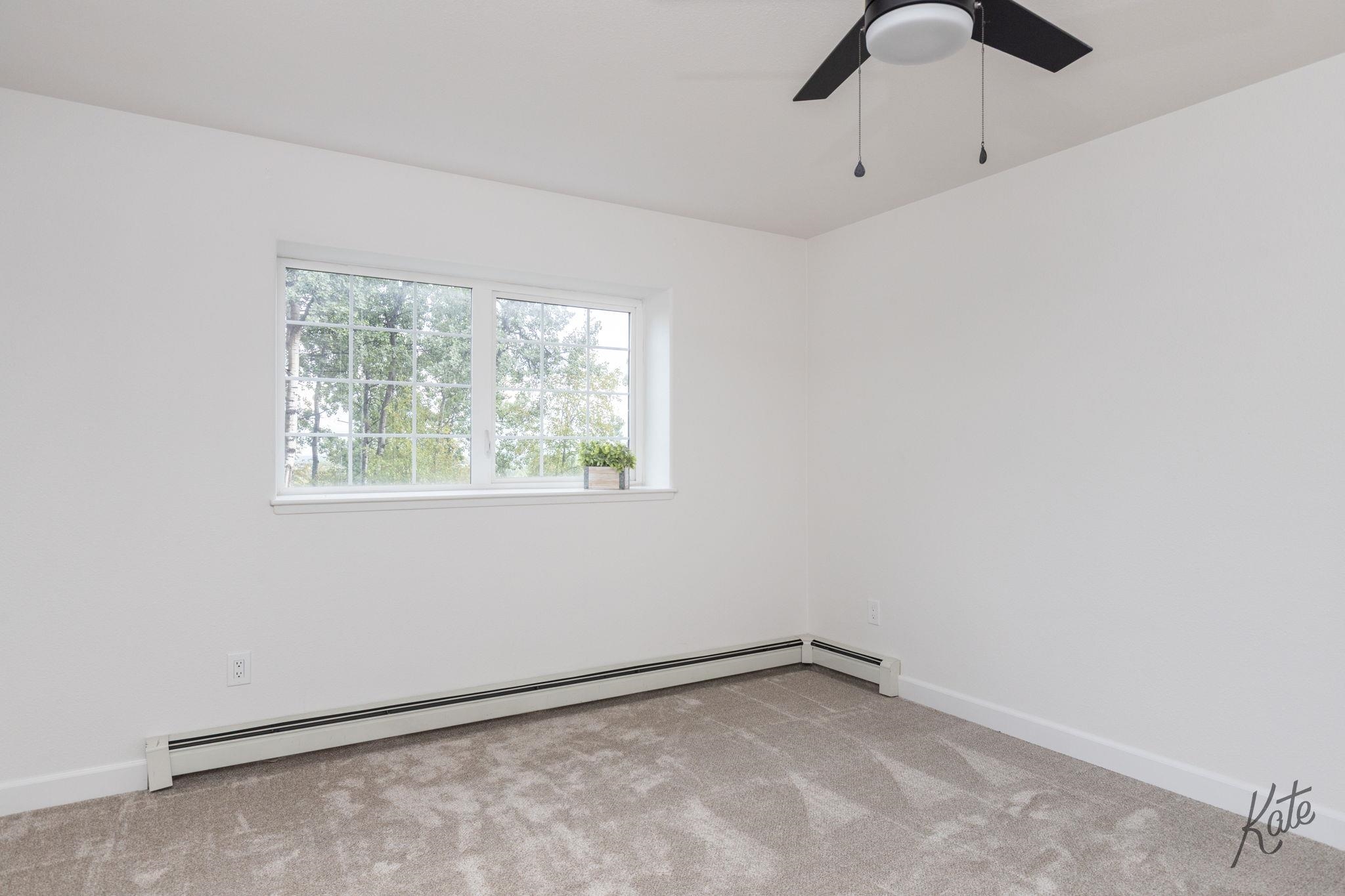 Empty carpeted room with a wide window, ceiling fan, and baseboard heater.