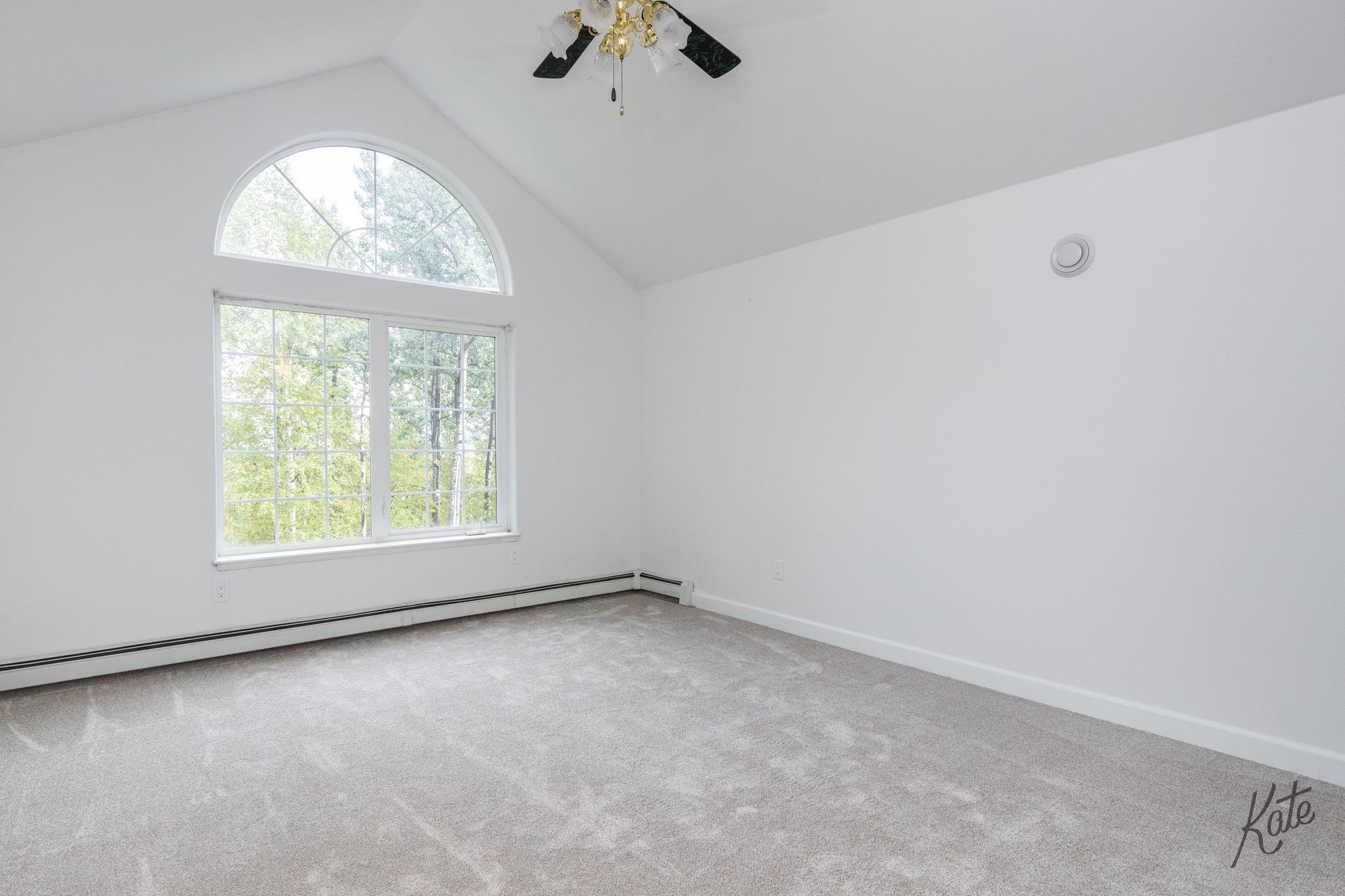 Empty bedroom with vaulted ceiling, gray carpet, and large arched window above a rectangular window.