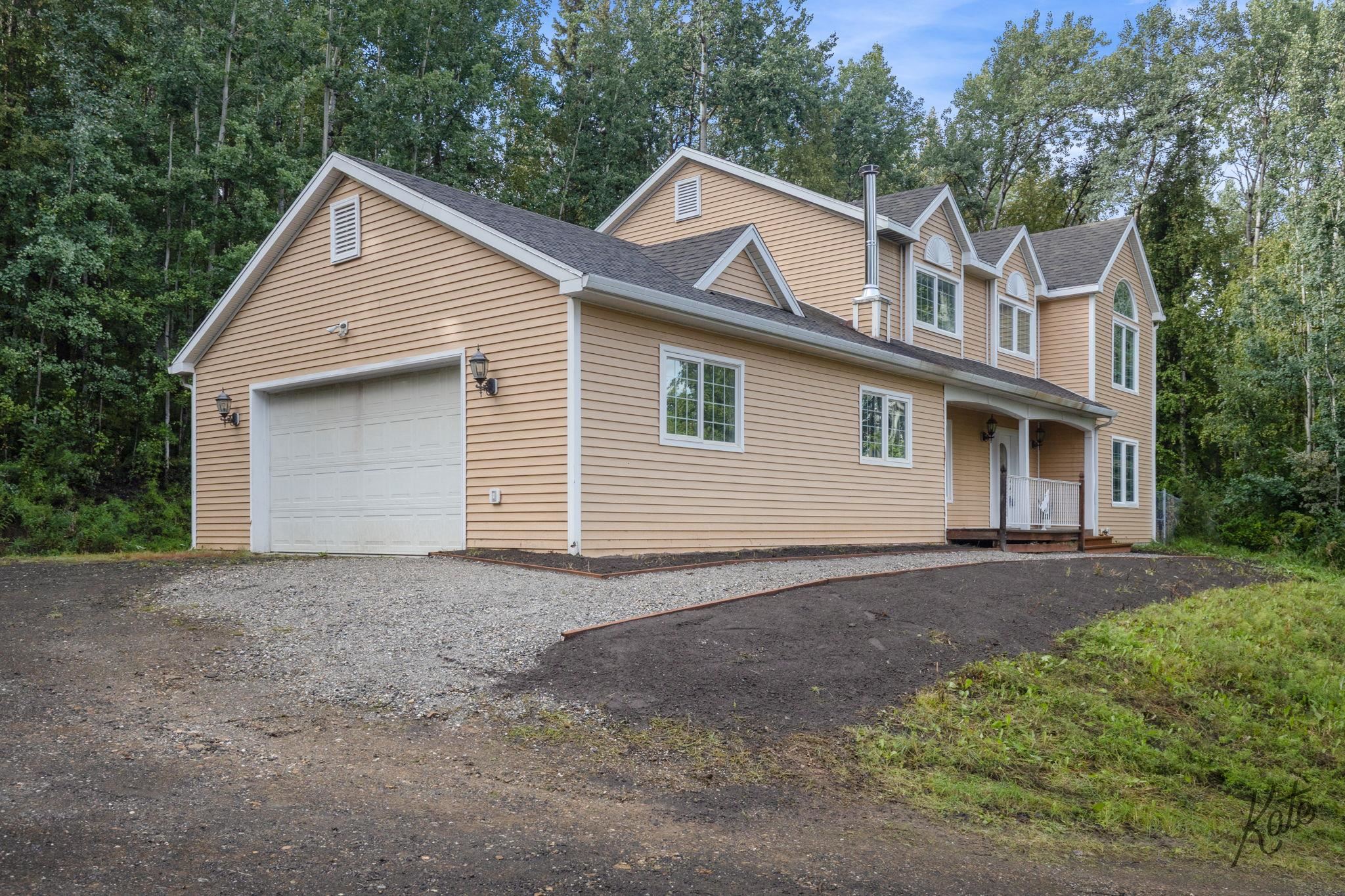 Tan two-story house with an attached garage, gravel driveway, and wooded surroundings.