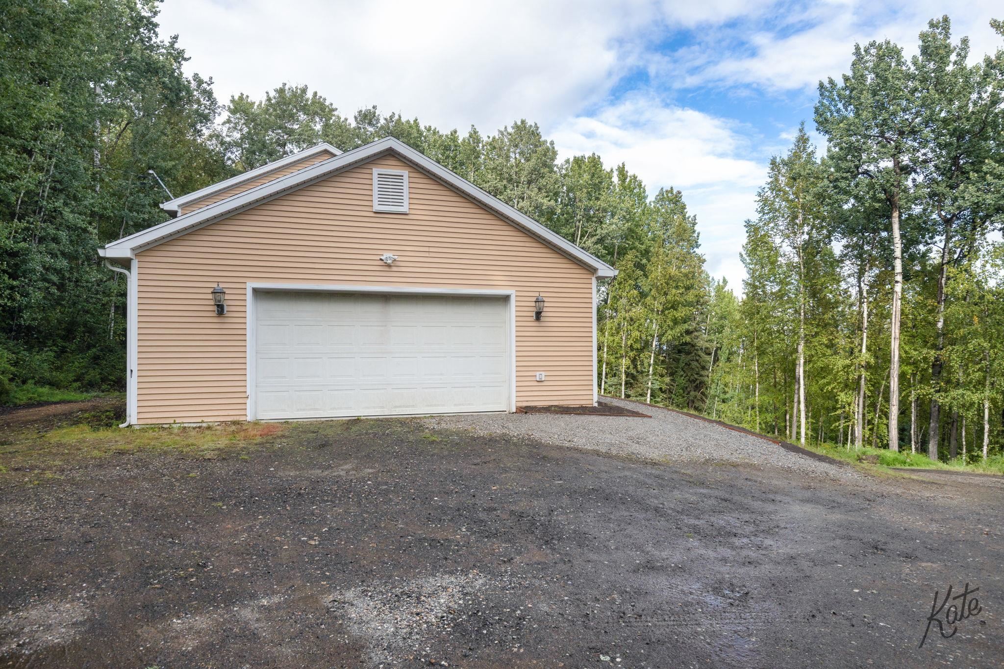 Tan detached garage with a white double door, gravel driveway, and surrounding trees.