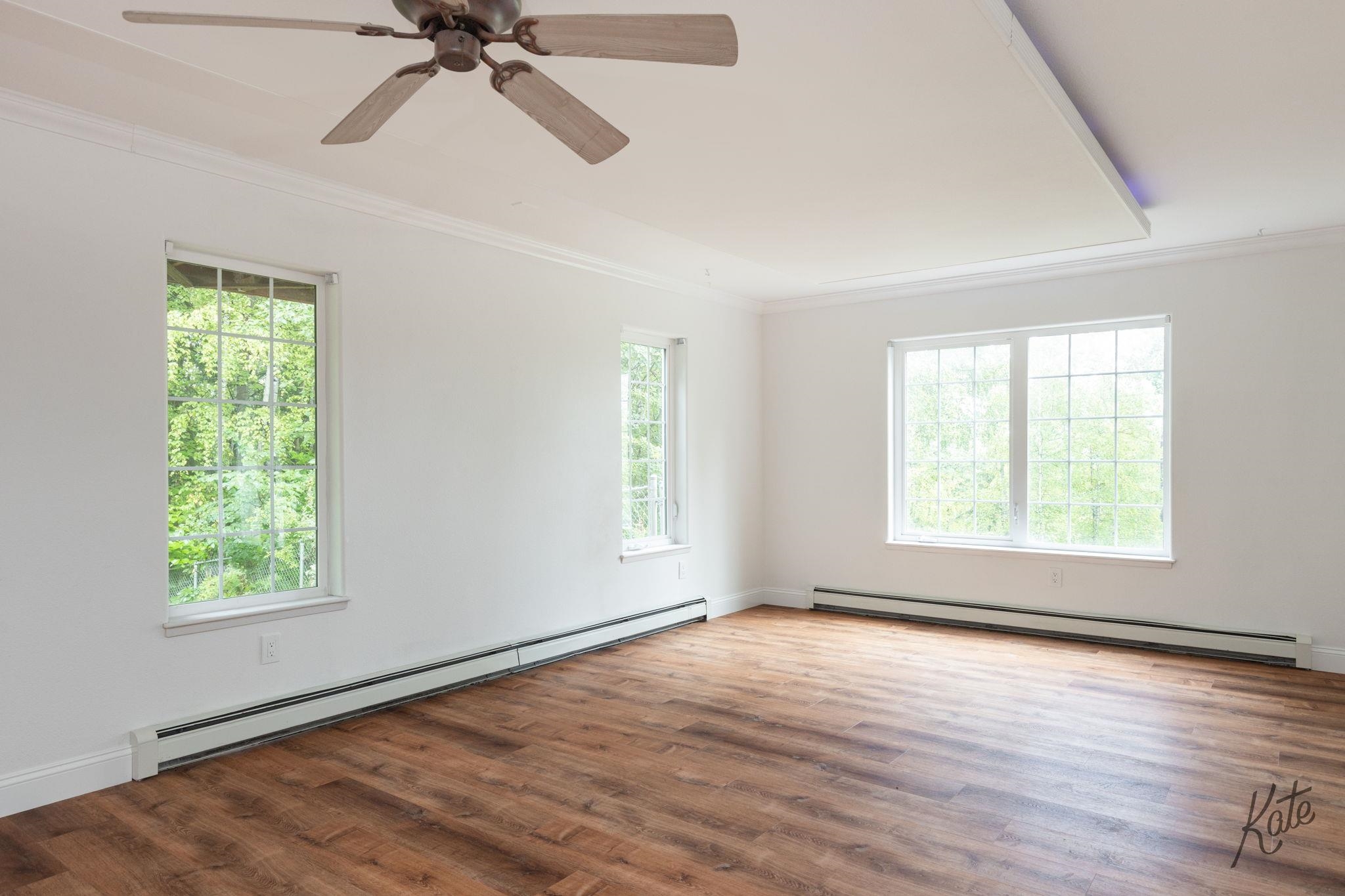 Empty room with wood floors, a ceiling fan, baseboard heaters, and large windows overlooking greenery.