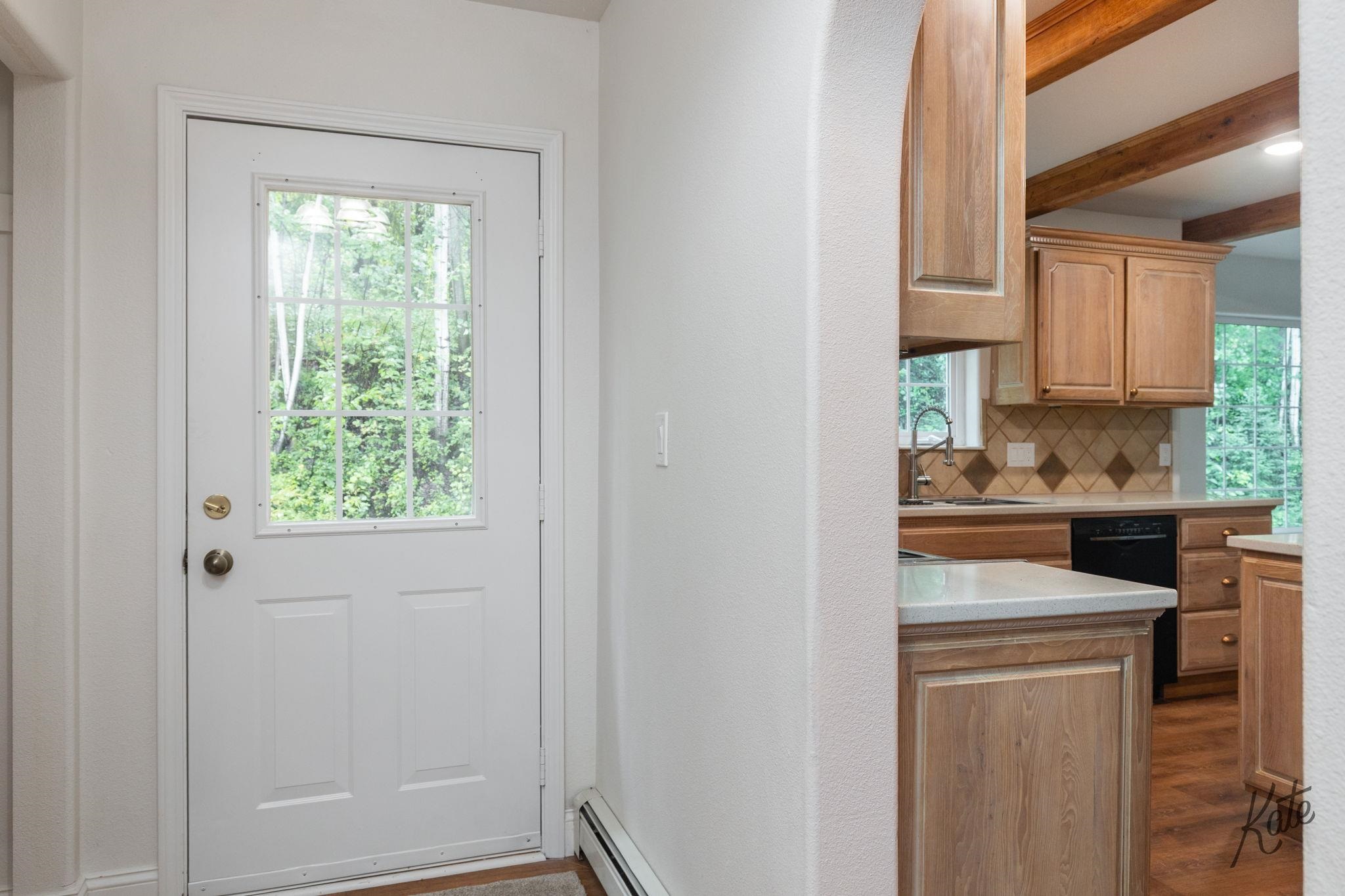 Entryway beside a wood-cabinet kitchen, with a windowed white door and exposed ceiling beams.